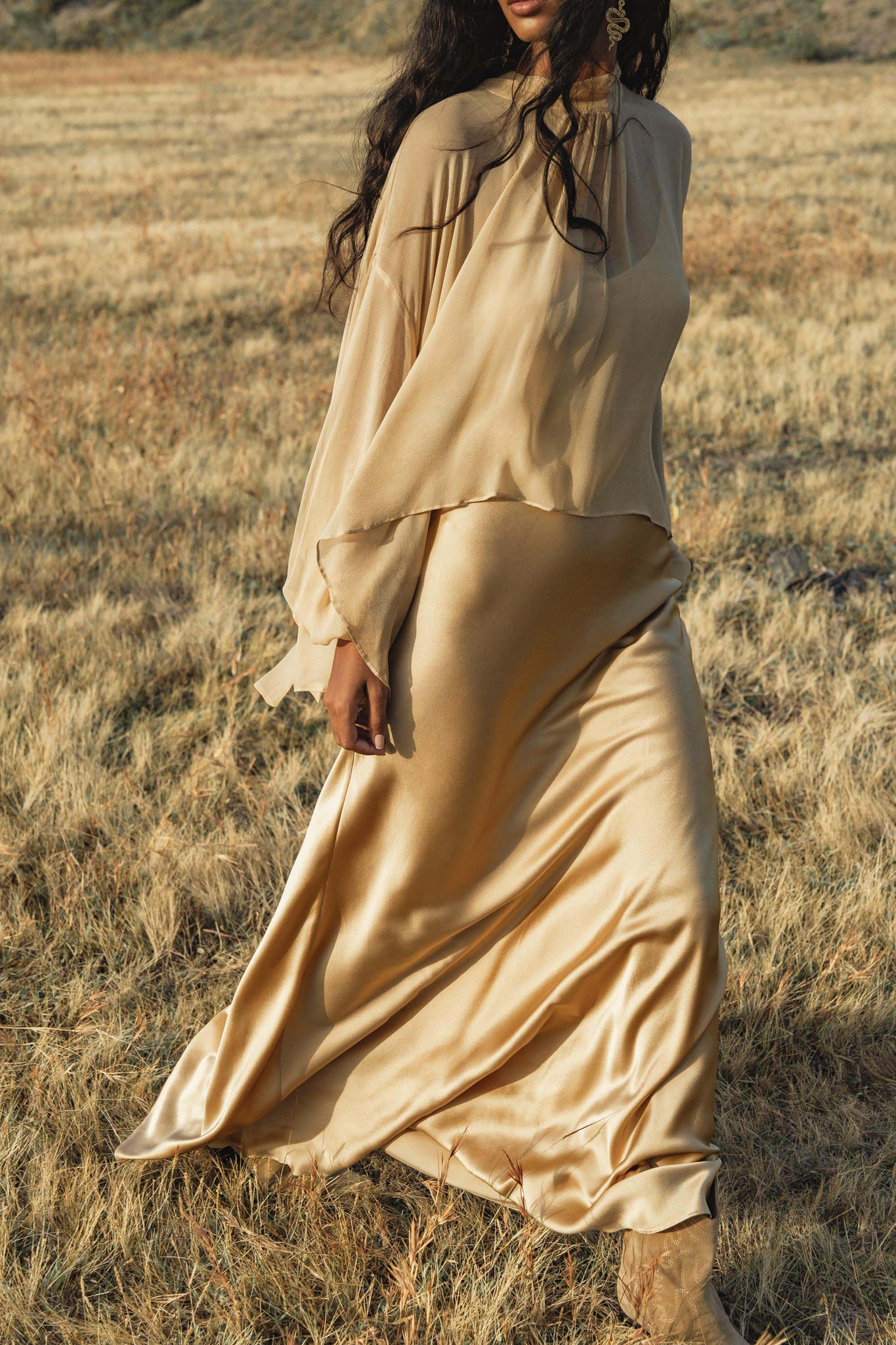 A woman wearing the AYA Sacred Wear Maharani Blouse in Inka Gold pure silk chiffon and matching skirt stands in a dry, grassy field, her face partially out of frame as her long dark hair cascades over her shoulders.