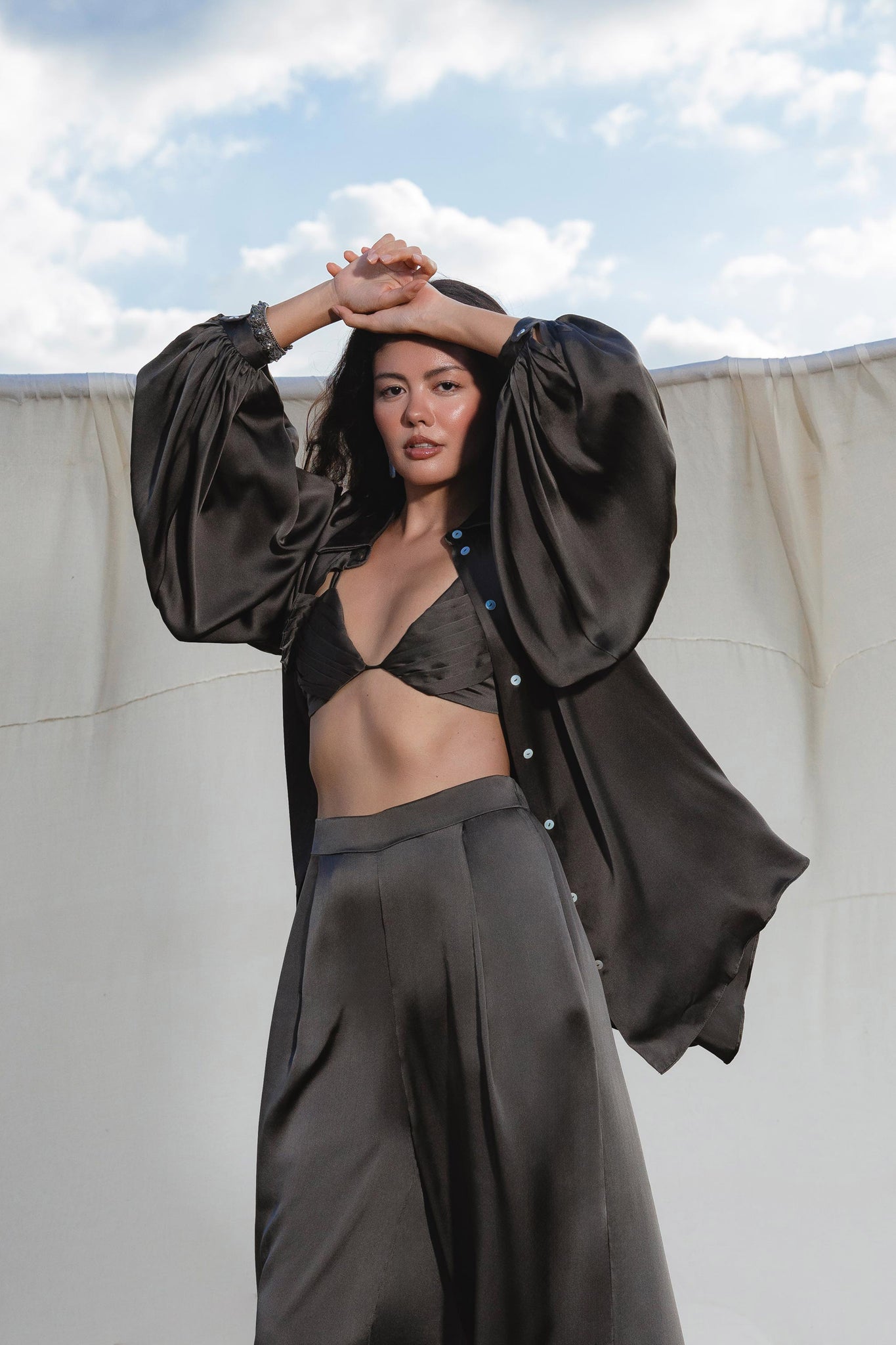 A woman poses outdoors before a sheer backdrop, wearing AYA Sacred Wear’s Apsara Pants in Asphalt Gray—pure silk, flowing silhouette trousers—paired with a black shirt and bralette. One arm is raised as she gazes at the camera.