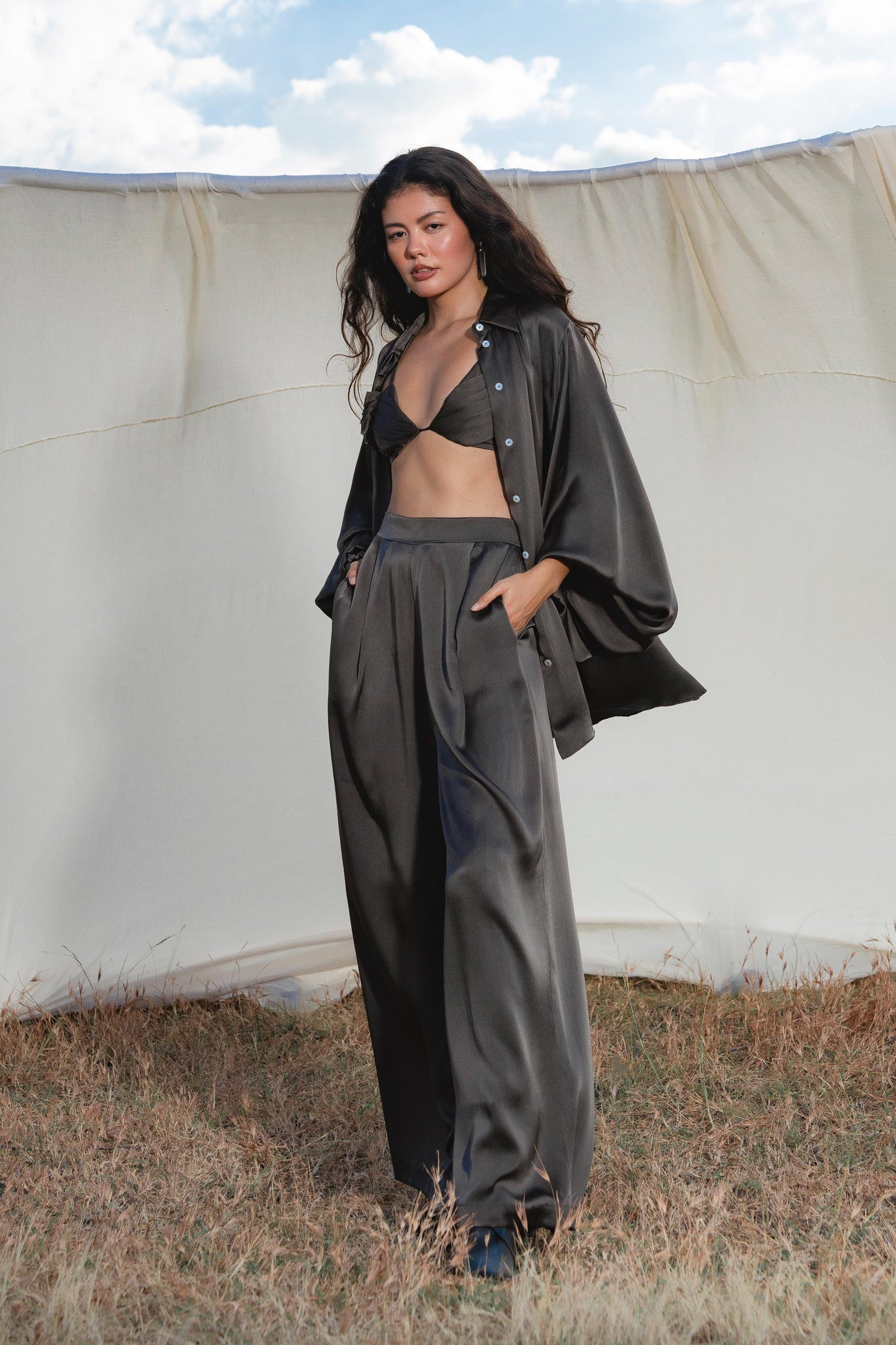 A woman with long dark hair stands on dry grass before a pale backdrop, wearing AYA Sacred Wear’s Apsara Pants in Asphalt Gray—pure silk, flowing silhouette trousers—with her hands in her pockets.