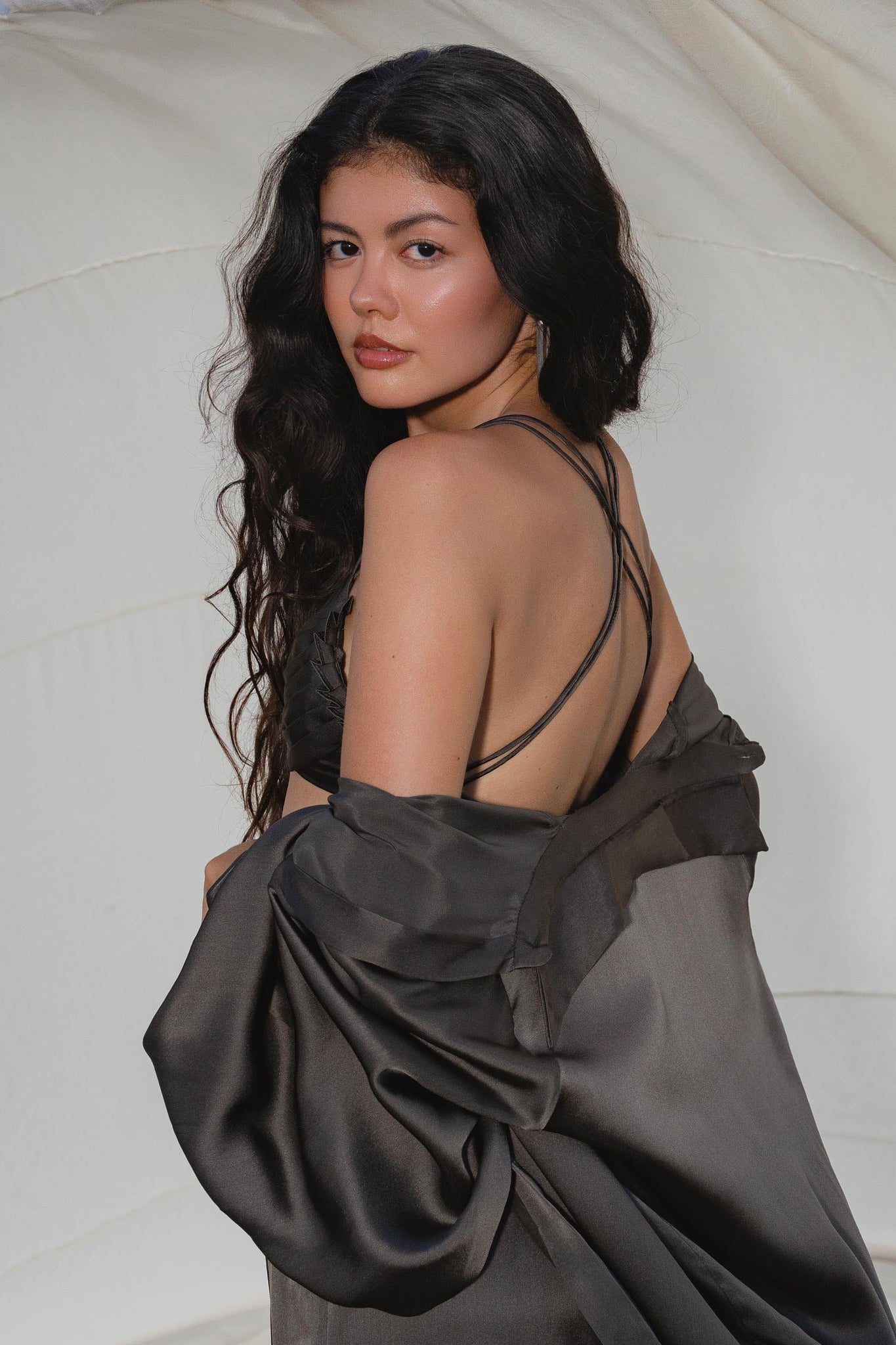 A woman with long, wavy dark hair wears the Butterfly Silk Bra in Asphalt Gray by AYA Sacred Wear—sculpted pure silk lingerie handcrafted in Bali. She stands against softly draped fabric, looking over her shoulder with a neutral expression.