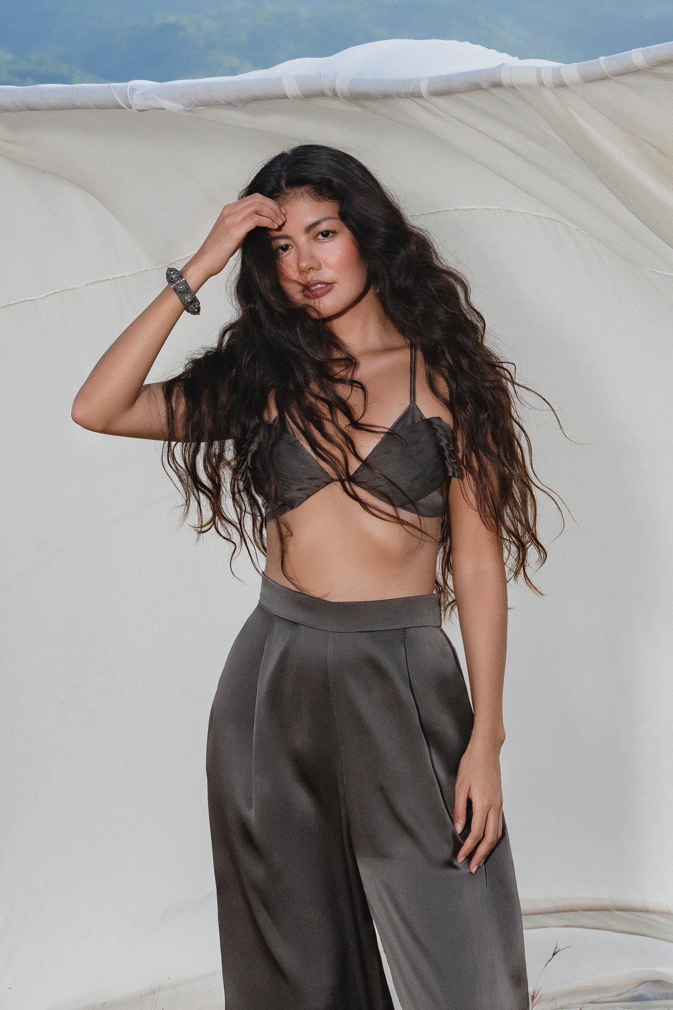 A woman with long wavy hair poses outdoors before a white fabric backdrop, wearing the Butterfly Silk Bra in Asphalt Gray from AYA Sacred Wear—sculpted pure silk lingerie—paired with matching high-waisted, wide-leg pants. She gazes at the camera.