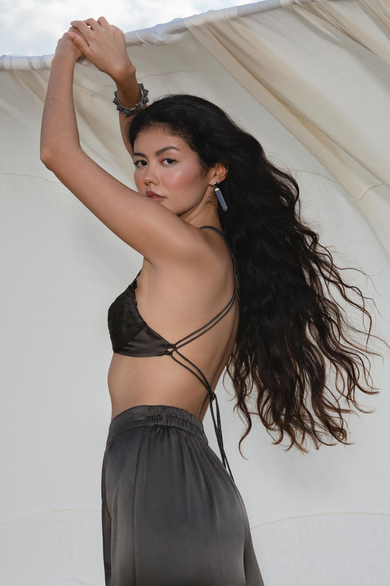 A woman with long, wavy dark hair poses outdoors in the Butterfly Silk Bra in Asphalt Gray by AYA Sacred Wear, standing before a light fabric backdrop and looking over her shoulder at the camera.