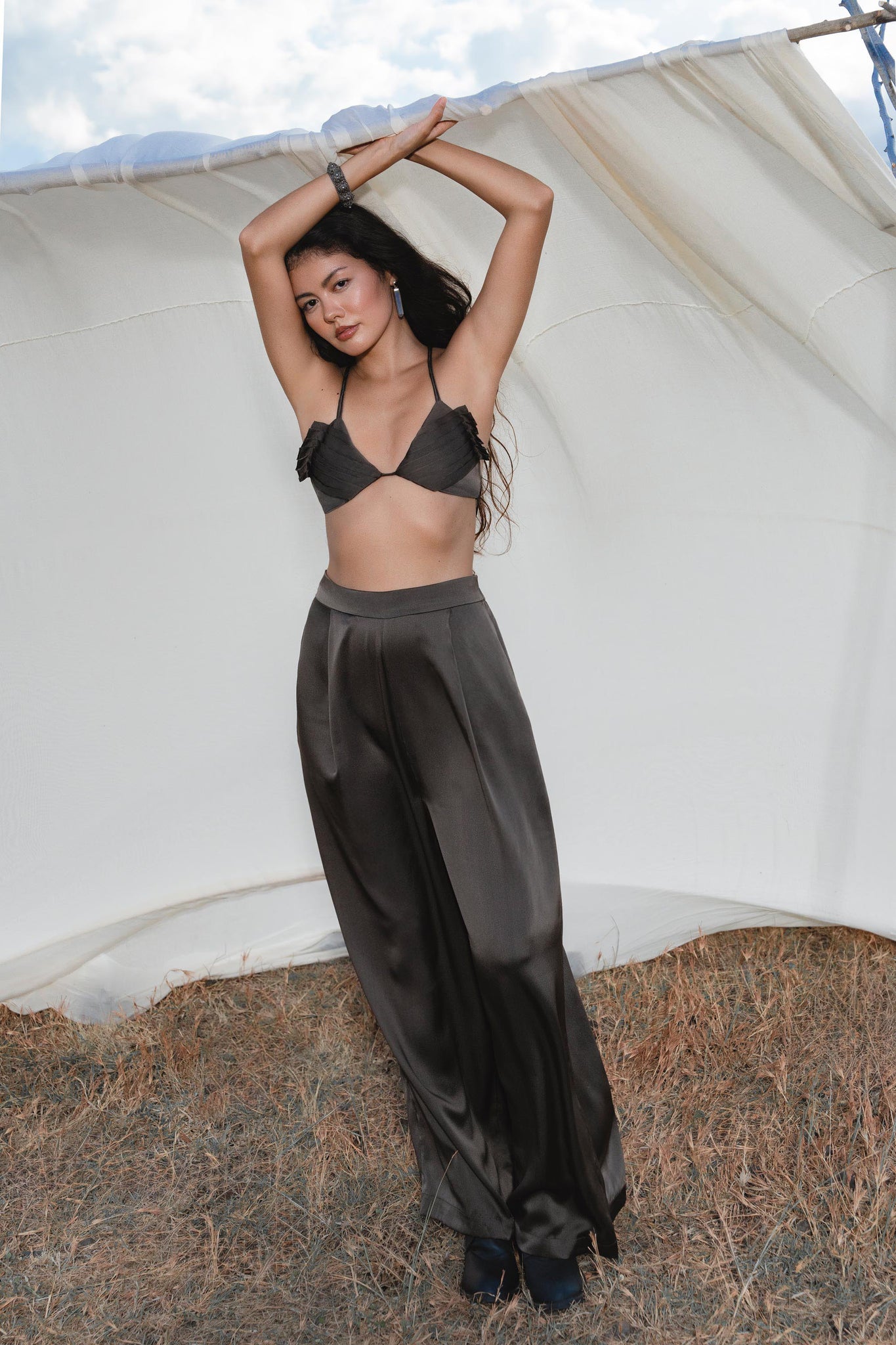 A woman in a black bralette and AYA Sacred Wear's Apsara Pants in Asphalt Gray — pure silk flowing silhouette trousers — stands on dry grass, arms raised, before a white fabric backdrop outdoors.