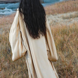A person with long, wavy dark hair models the AYA Sacred Wear Gold Silk Long Robe—a floor-length, relaxed bohemian kimono—while standing in a dry, grassy field near a river, facing away from the camera.