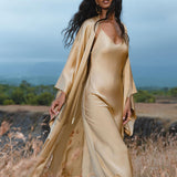 A woman in the AYA Sacred Wear Gold Silk Long Robe— a floor-length maxi kimono and relaxed bohemian cardigan—stands in a grassy field, her long dark hair and gold earrings shining against trees and a cloudy sky.