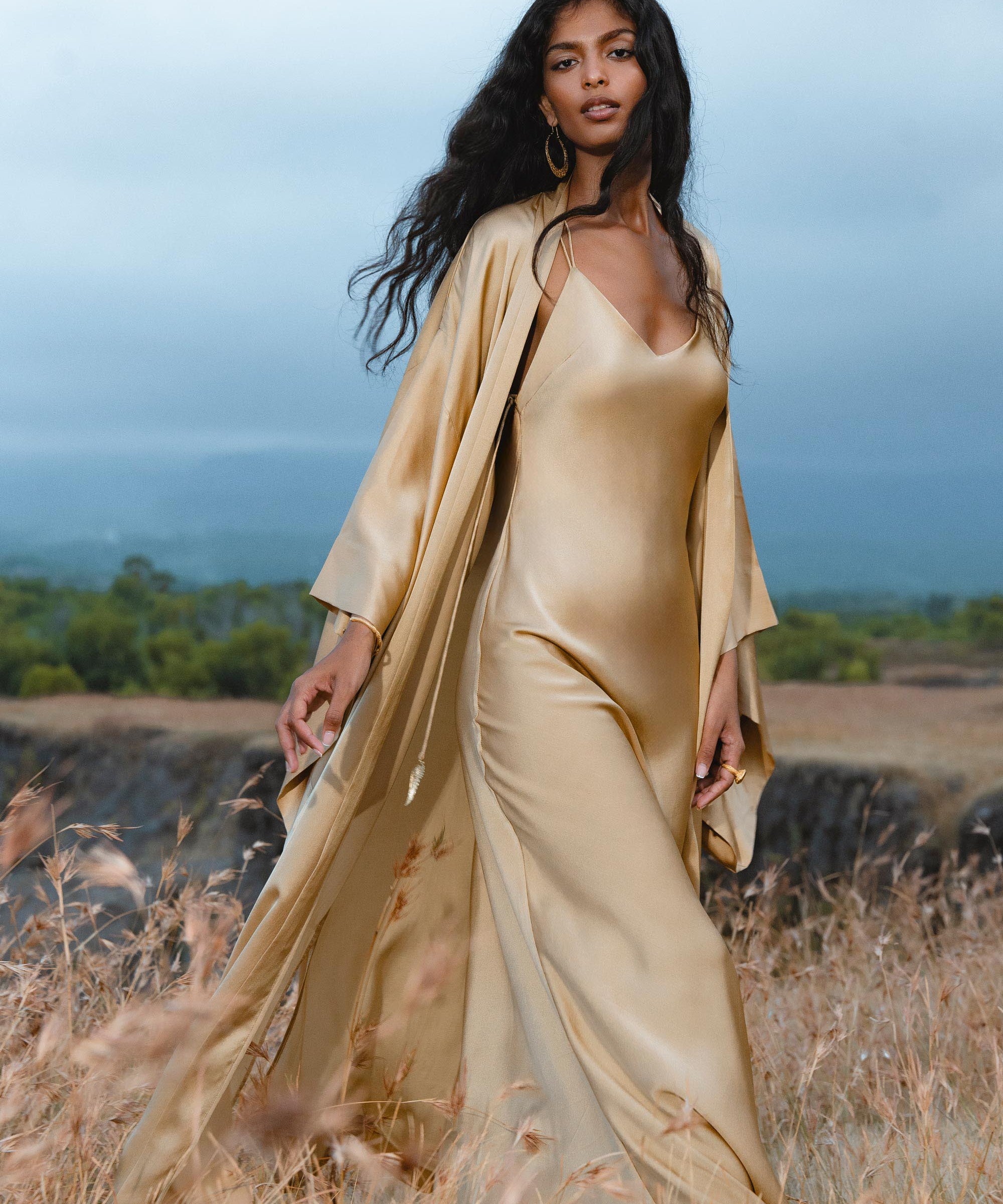 A woman in the AYA Sacred Wear Gold Silk Long Robe— a floor-length maxi kimono and relaxed bohemian cardigan—stands in a grassy field, her long dark hair and gold earrings shining against trees and a cloudy sky.
