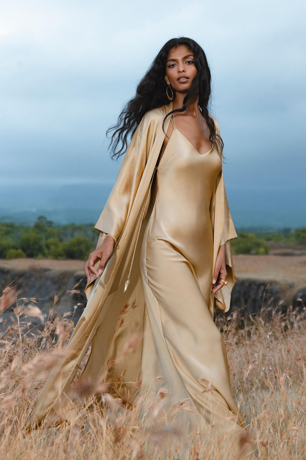 A woman in the AYA Sacred Wear Gold Silk Long Robe— a floor-length maxi kimono and relaxed bohemian cardigan—stands in a grassy field, her long dark hair and gold earrings shining against trees and a cloudy sky.