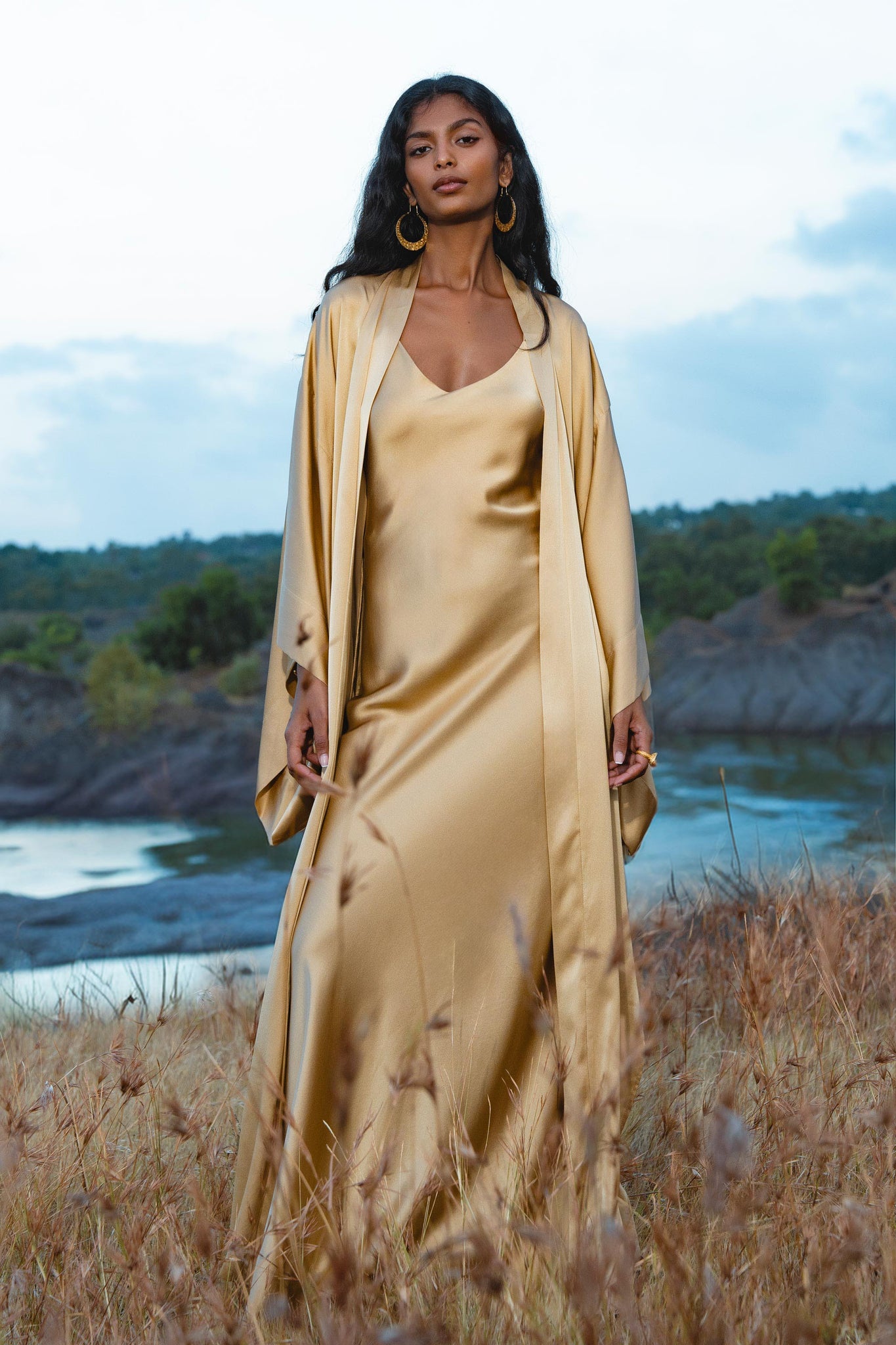 A woman with long dark hair and gold hoop earrings stands outdoors in tall grass, wearing the Gold Silk Long Robe— a floor-length maxi kimono by AYA Sacred Wear—against a serene backdrop of water, hills, and a cloudy sky.