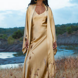 A woman with long dark hair and gold hoop earrings stands outdoors in tall grass, wearing the Gold Silk Long Robe— a floor-length maxi kimono by AYA Sacred Wear—against a serene backdrop of water, hills, and a cloudy sky.