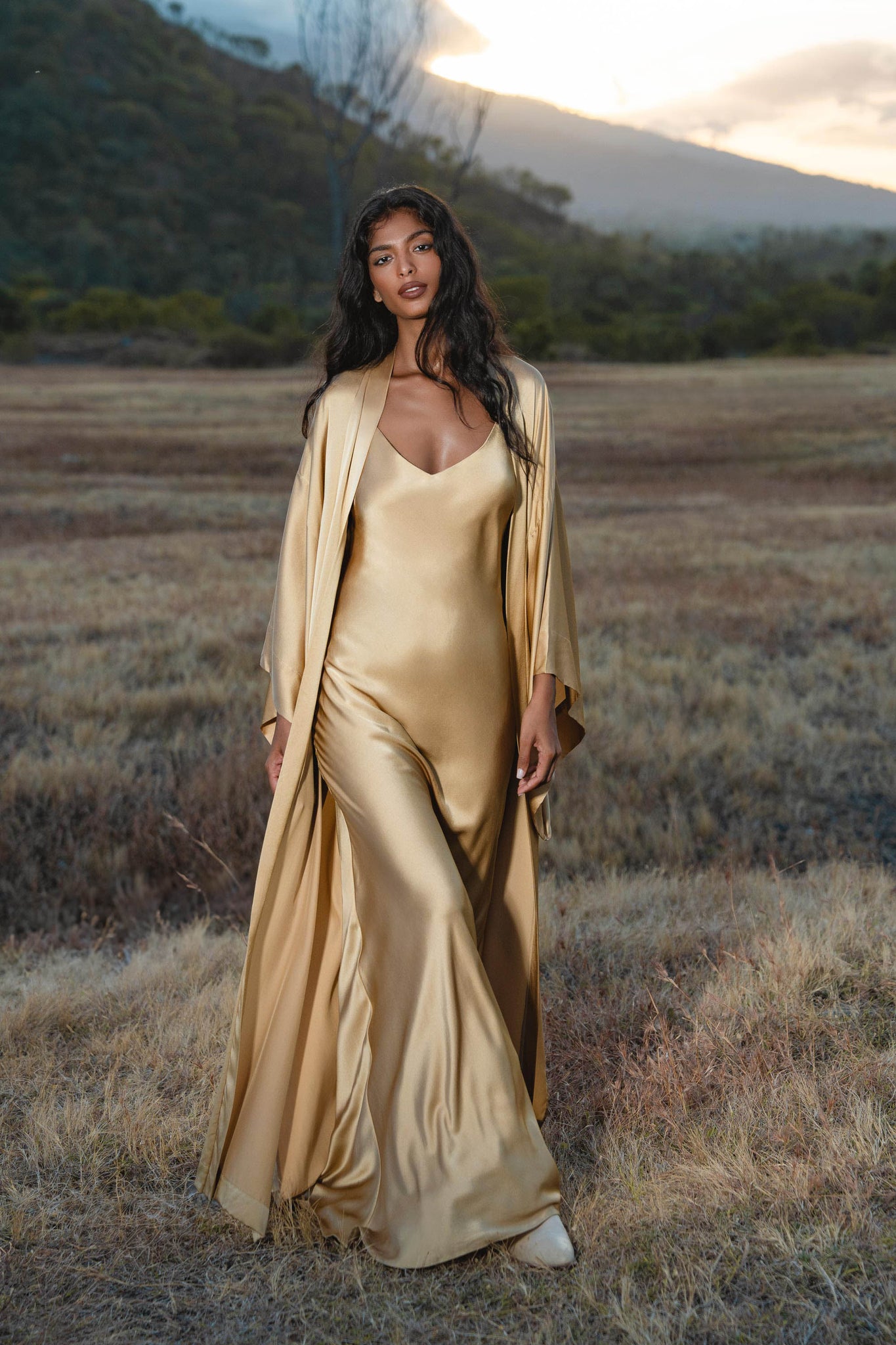 At sunset, a woman stands in a dry grassy field wearing the AYA Sacred Wear Gold Silk Long Robe — a floor-length maxi kimono and relaxed bohemian cardigan. Mountains and trees are visible beneath the partly cloudy sky in the background.
