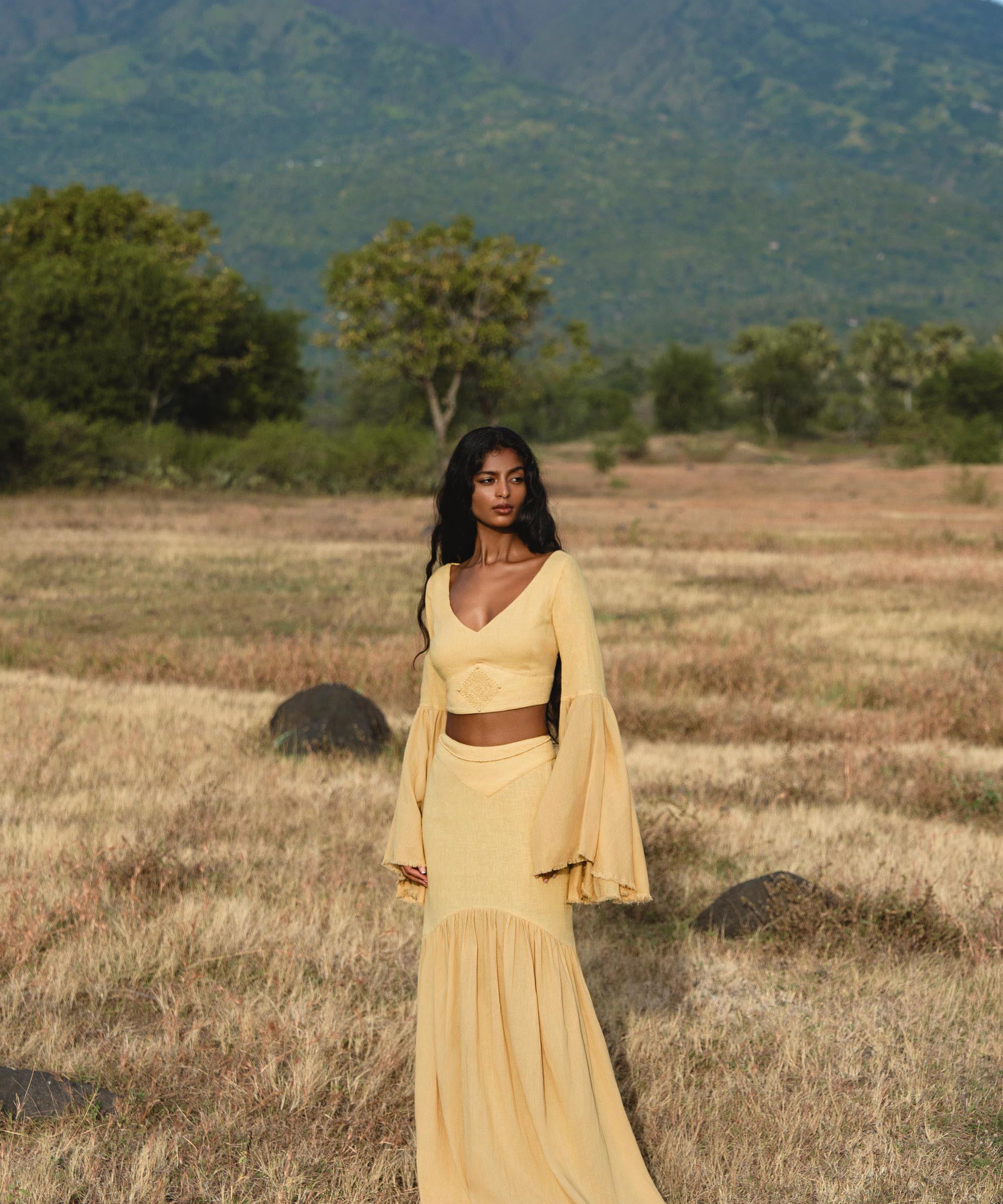 A woman wears the AYA Sacred Wear Indian Set in Yellow Gold—a cotton two-piece outfit with a flowy bell-sleeve top and maxi skirt—standing in a dry grassy field with green trees and mountains behind her under a blue sky.