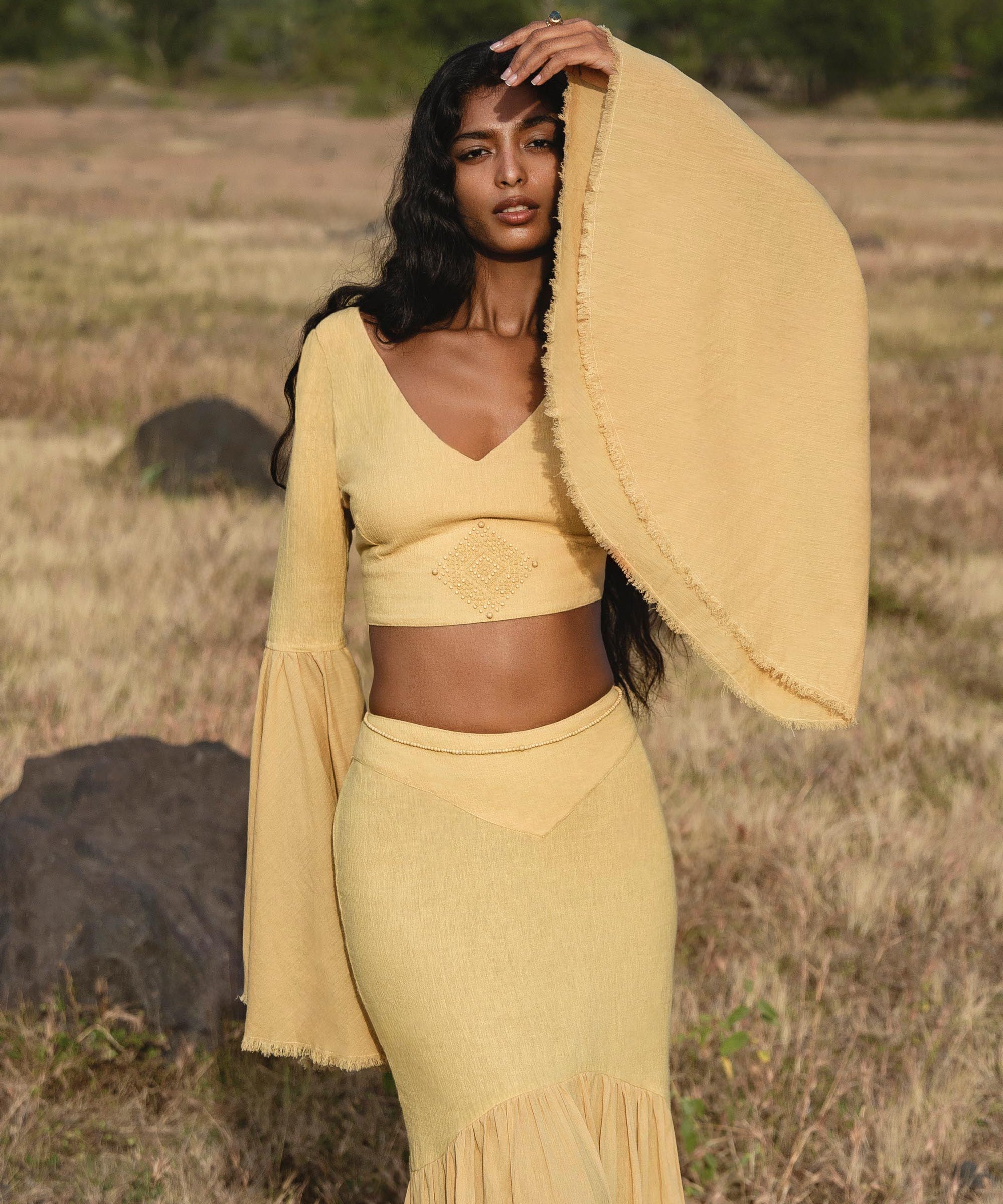 A woman wears the AYA Sacred Wear Indian Set in Yellow Gold—a flowy bell-sleeve cotton crop top and maxi skirt—in a dry grassy field, with trees and bushes in the background and her hand raised to her forehead.