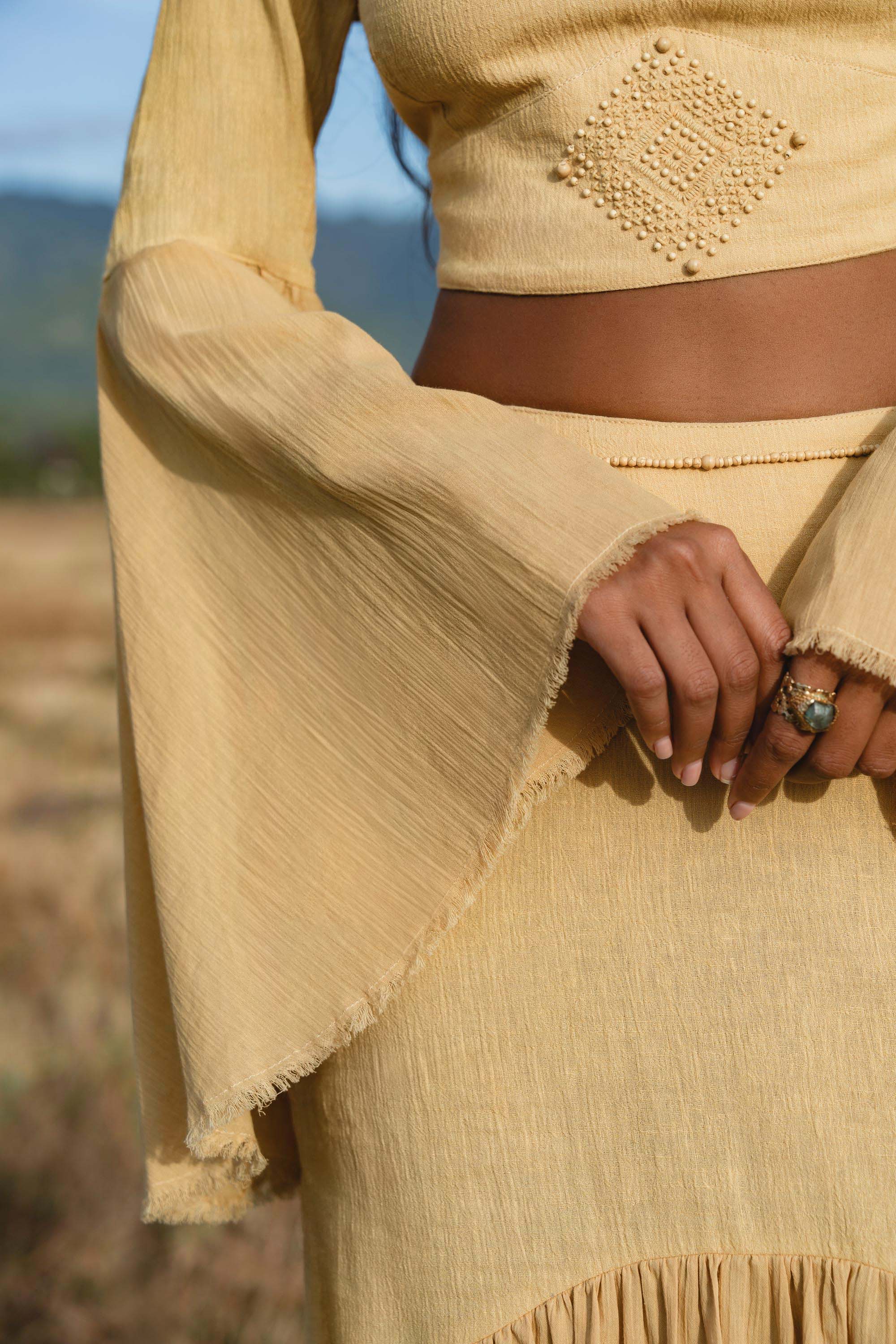 In a sunlit field, a person models AYA Sacred Wear’s Indian Set in Yellow Gold—a cotton two-piece with a flowy bell-sleeve top and maxi skirt. The geometric pattern and breathable fabric give this outfit effortless charm.