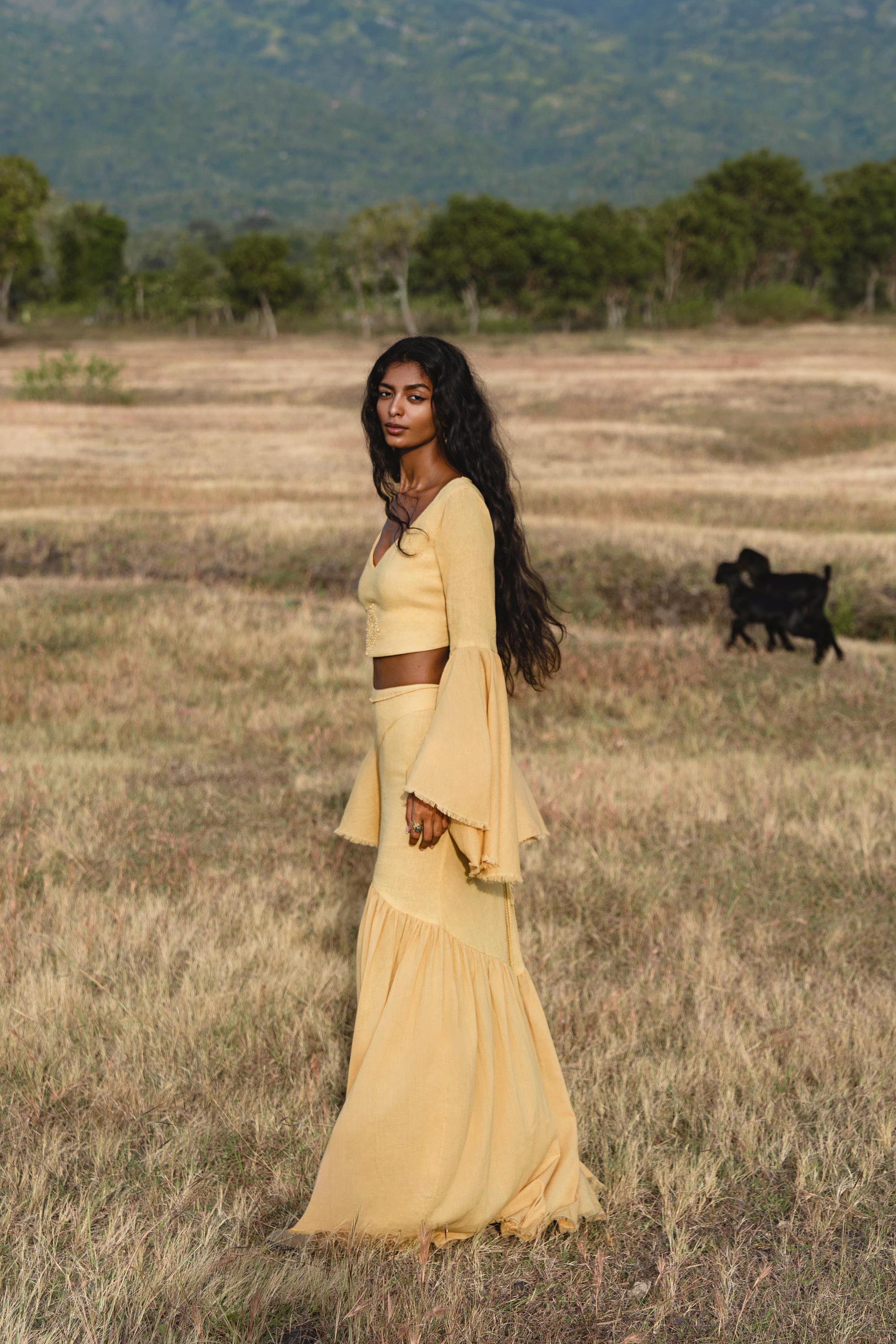 A woman wearing the AYA Sacred Wear Indian Set in Yellow Gold—cotton two-piece with flowy bell-sleeve top and maxi skirt—stands in a dry grassy field with trees, mountains, and a black dog in the background.