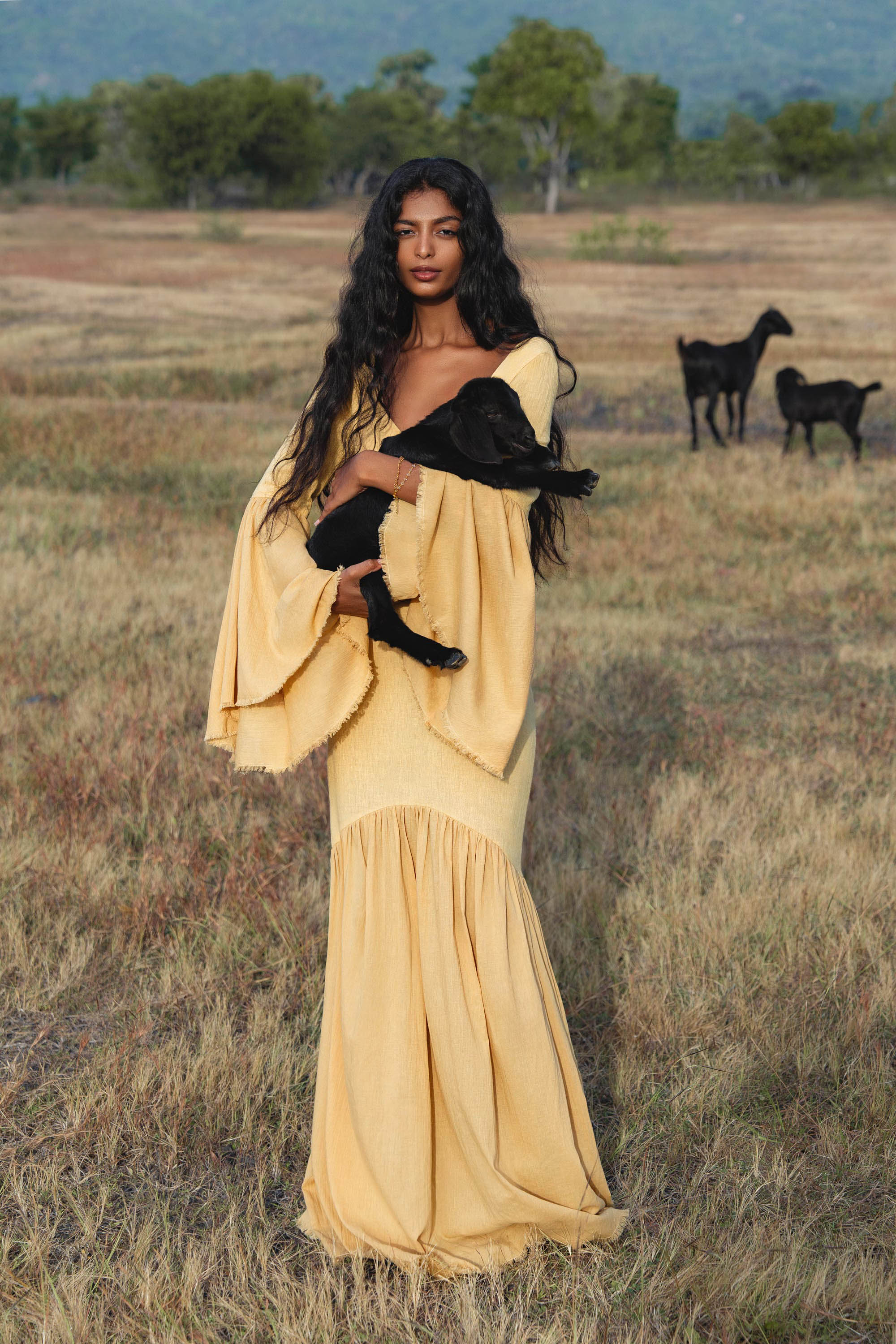 A woman in the AYA Sacred Wear Indian Set in Yellow Gold—a cotton two-piece with a flowy bell-sleeve top and maxi skirt—stands in a grassy field holding a black baby goat, with two adult goats grazing nearby and hills beyond.