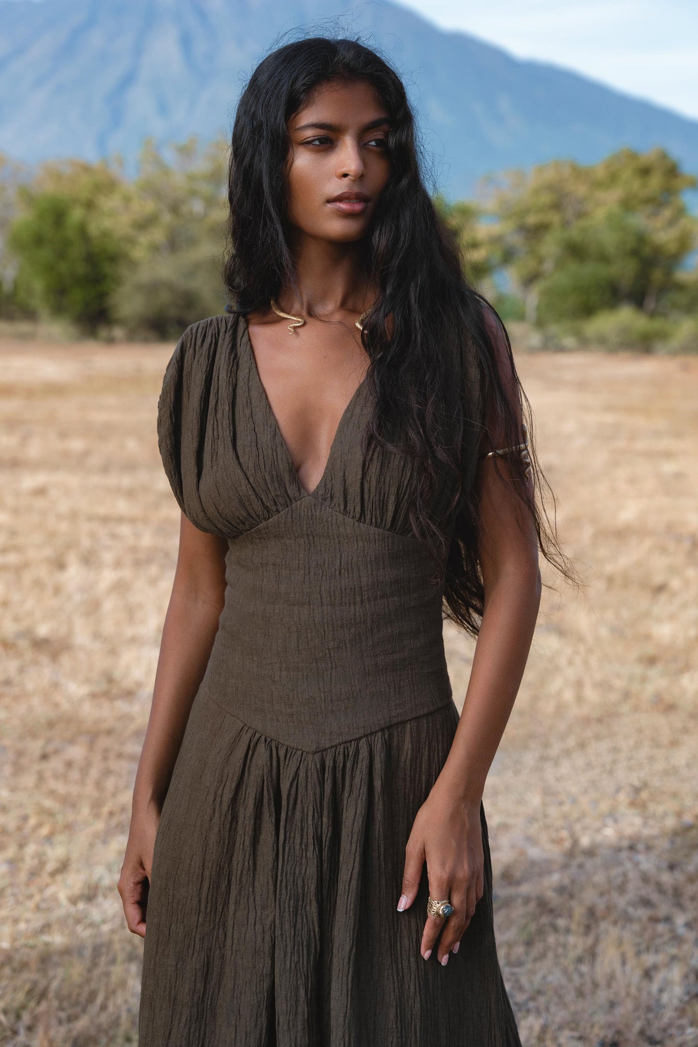 A woman with long dark hair wears the Japan Dress in African Brown by AYA Sacred Wear, a cotton maxi dress with gathered shoulders and a flowing skirt, standing outdoors in a dry grassy field with trees and mountains behind her.