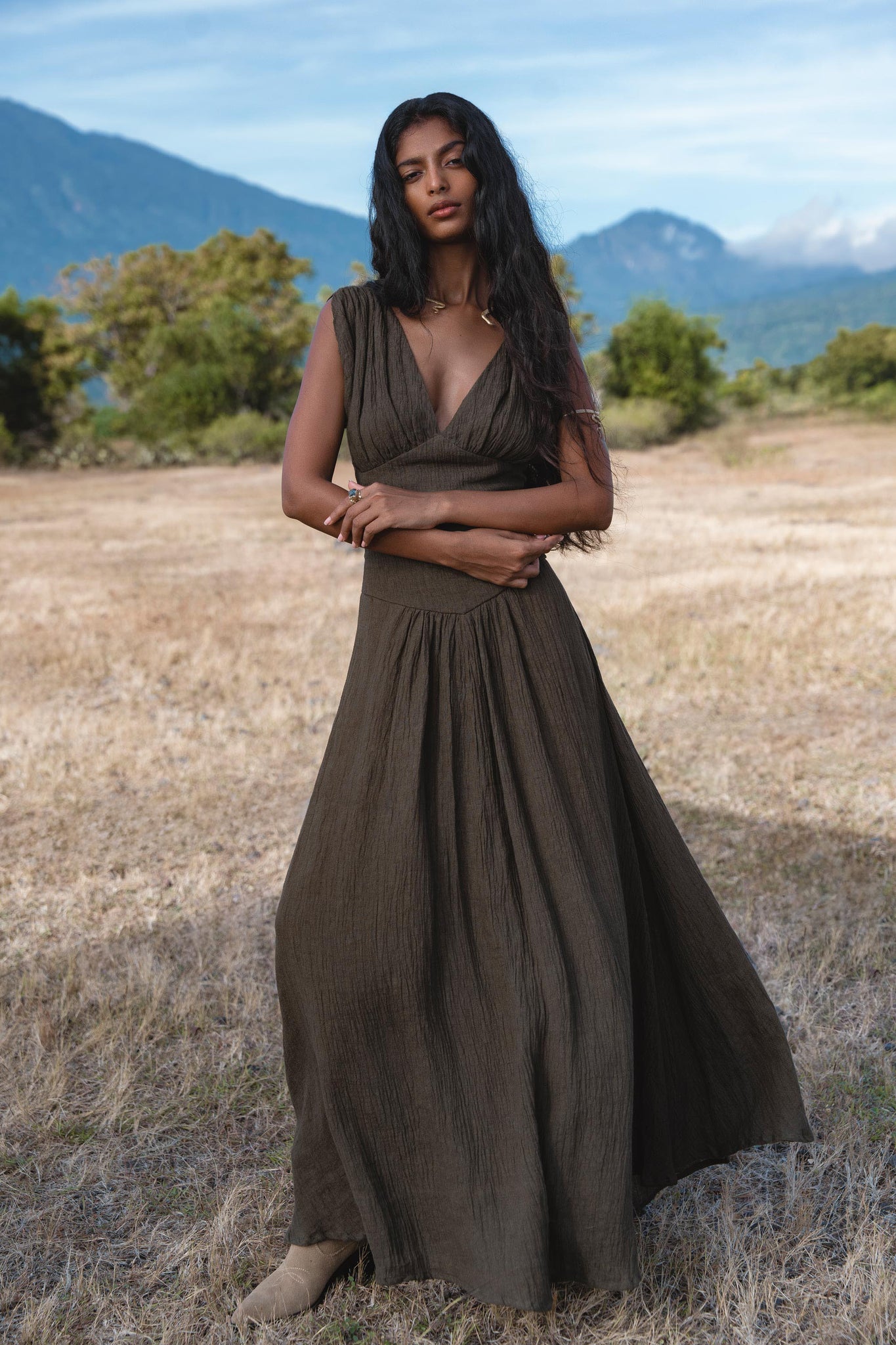 A woman with long dark hair stands in a dry, grassy field wearing the AYA Sacred Wear Japan Dress in African Brown—a cotton maxi dress with gathered shoulders and a flowing skirt. Mountains and trees are seen under a blue sky.