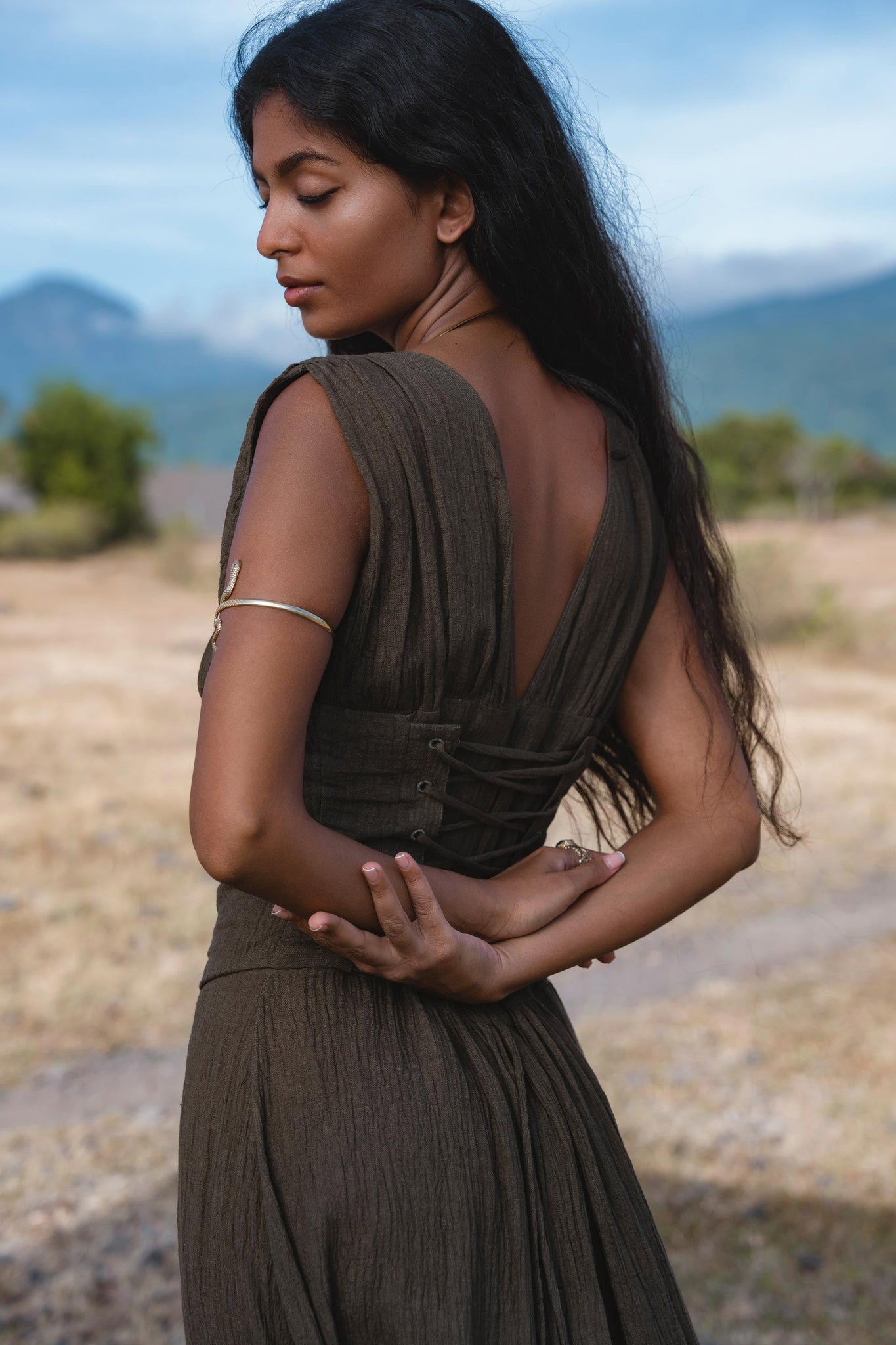 A woman with long dark hair stands outdoors, looking down with arms crossed behind her back, wearing the AYA Sacred Wear Japan Dress in African Brown—a cotton maxi dress with gathered shoulders and a flowing skirt. Mountains and dry grassland are behind her.