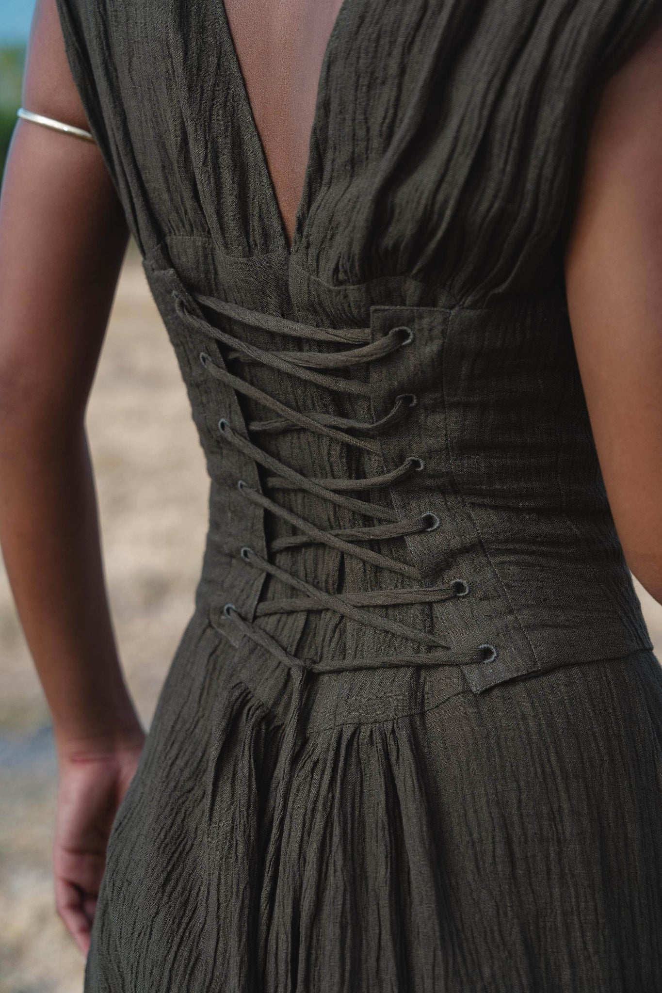 Close-up of a person in the Japan Dress in African Brown by AYA Sacred Wear, featuring a gathered shoulder silhouette and flowing cotton maxi skirt, with a thin bangle accent and an elegant, softly focused outdoor backdrop.