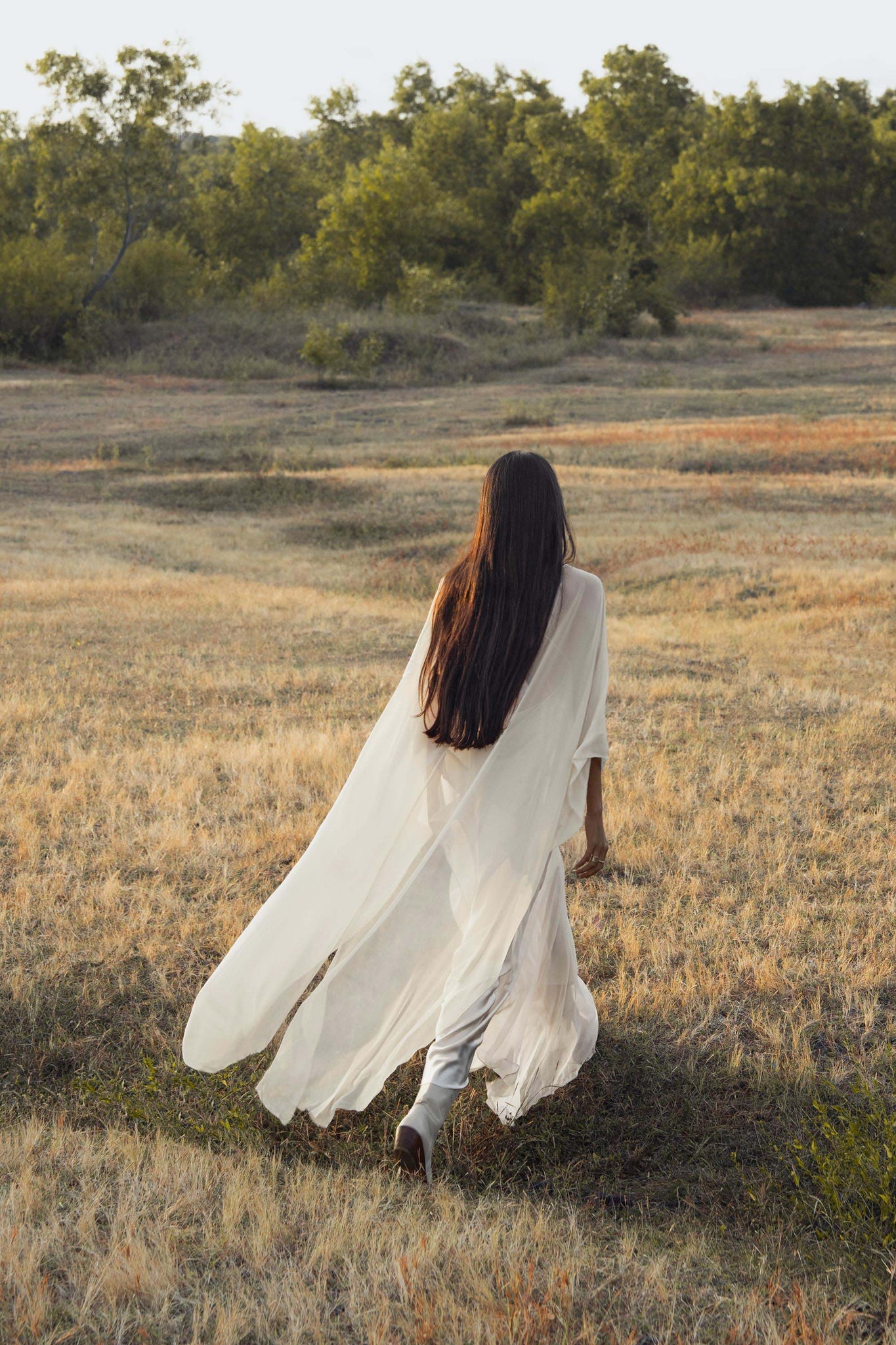 A woman with long dark hair, wearing the Mahadevi Dress Cream Colour – Pure Silk Chiffon Goddess Gown by AYA Sacred Wear, walks across a dry grassy field toward distant green trees in gentle natural light.