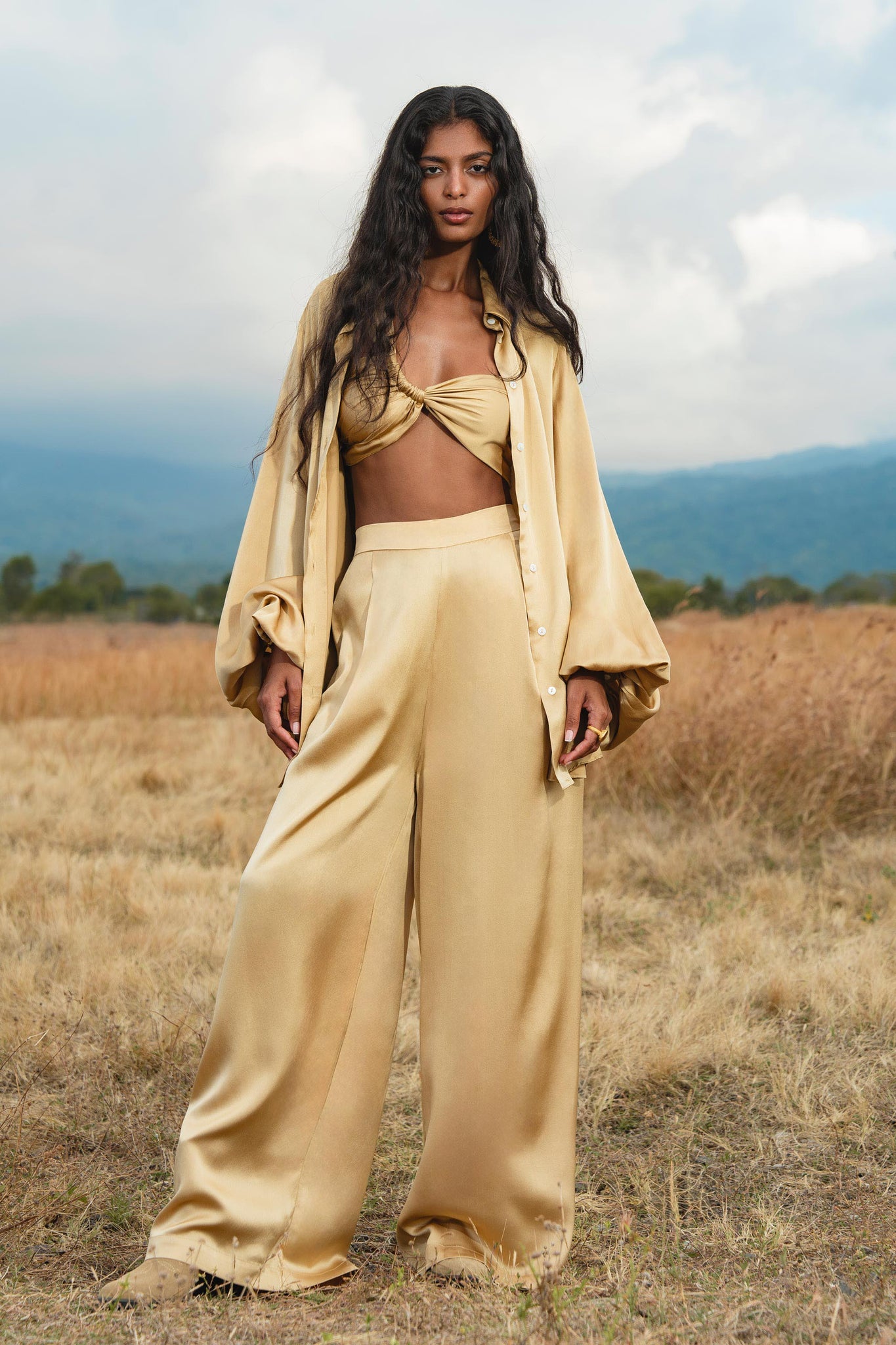 A woman stands in a dry grassy field wearing the Apsara Bra Inka Gold Colour—Pure Silk Transformative Top by AYA Sacred Wear, with matching gold satin pieces, as mountains and a cloudy sky form the background.