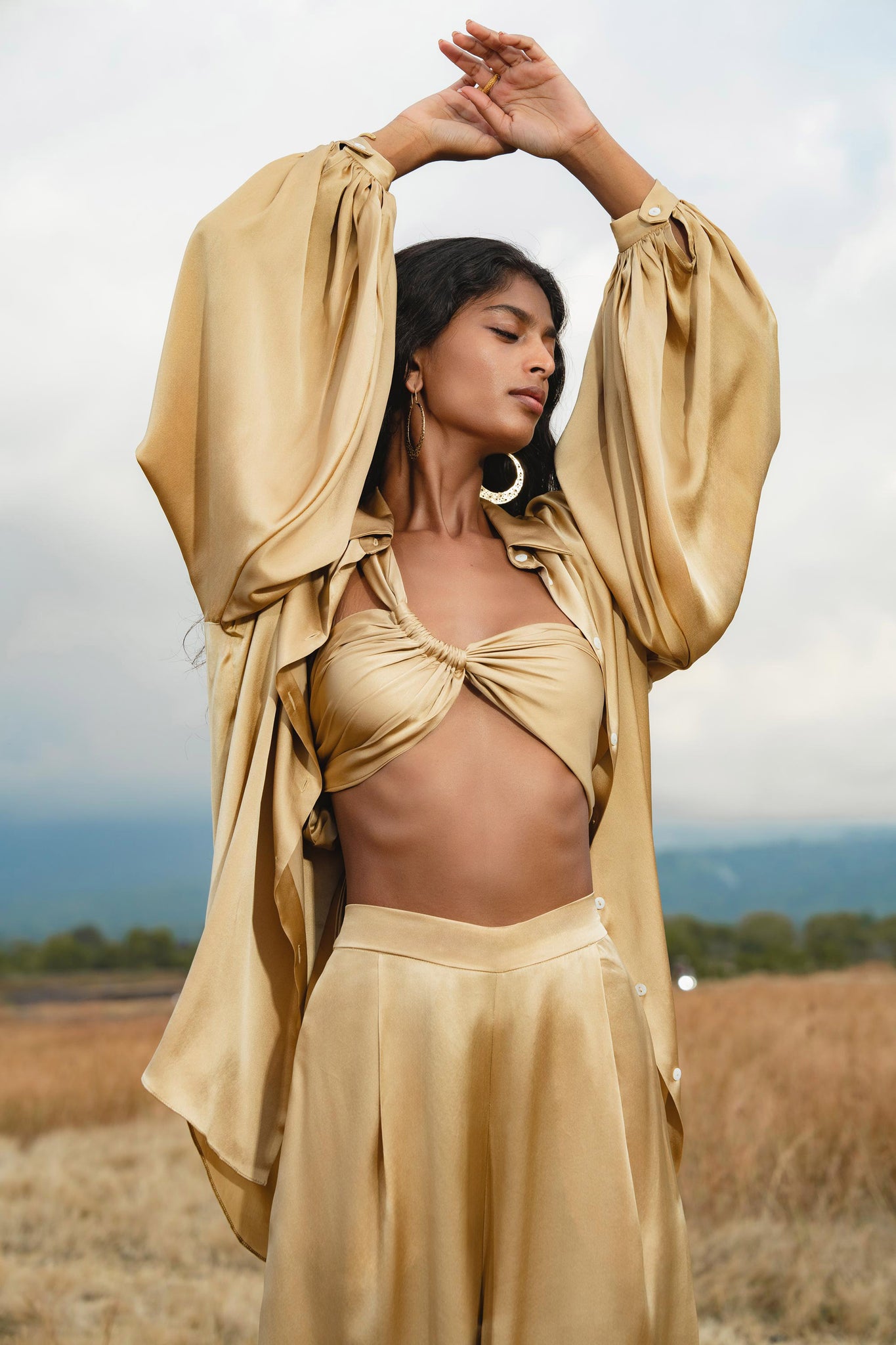 A woman stands outdoors in a field, wearing the AYA Sacred Wear Apsara Kimono Blouse Inka Gold Colour—an elegant, pure silk kimono top handcrafted in Bali—posing with arms raised under a cloudy sky with trees and hills behind her.