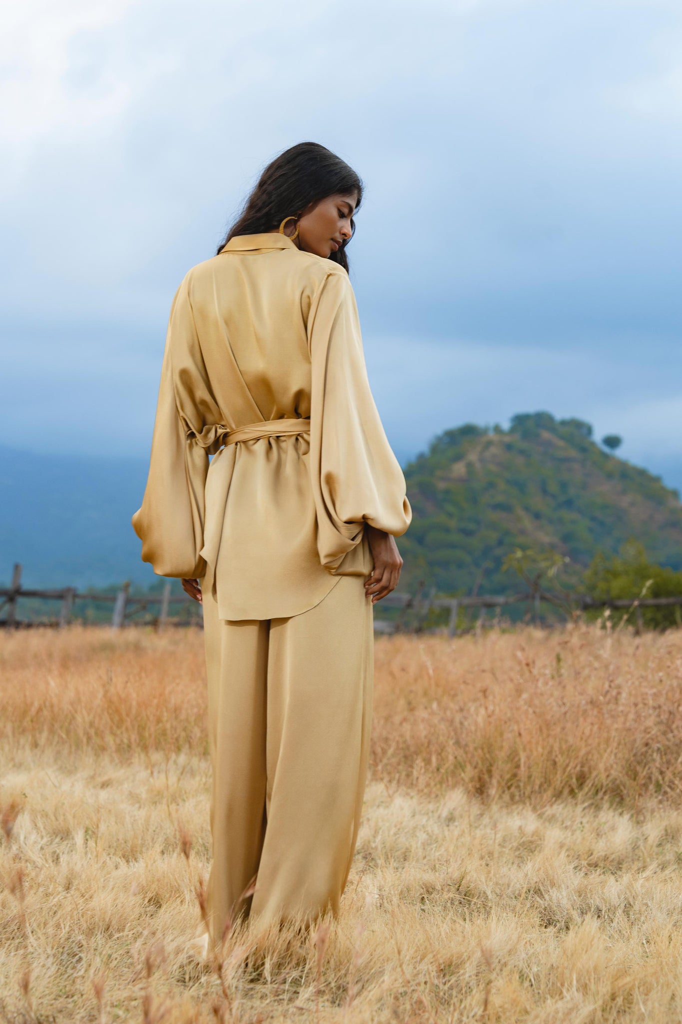 A woman stands in a grassy field with hills behind her, wearing AYA Sacred Wear's Apsara Pants Inka Gold—pure silk flowing silhouette trousers—in gold. She looks over her shoulder as clouds fill the sky.