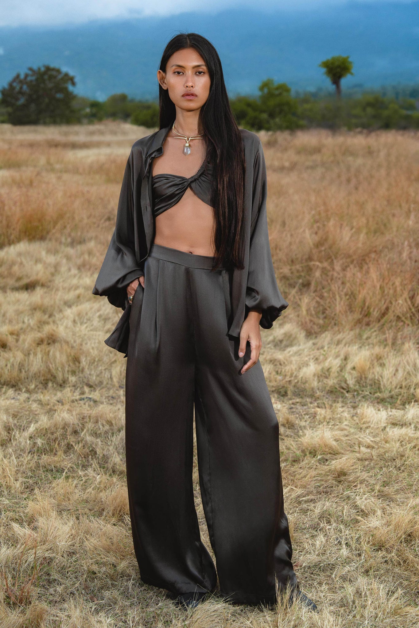 A person with long dark hair stands in a dry grassy field wearing the Apsara Kimono Blouse in Asphalt Gray by AYA Sacred Wear, handcrafted from pure silk. Mountains and trees are visible in the background.
