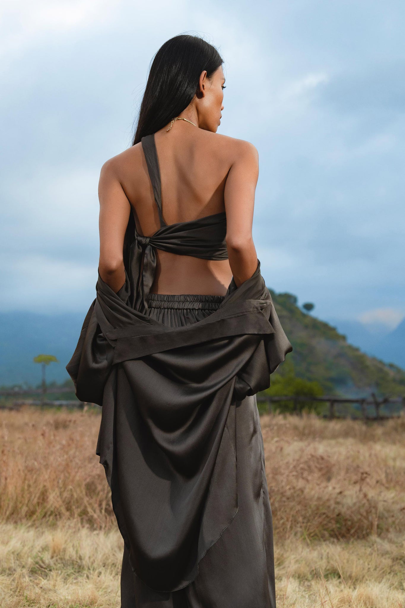 A woman with long dark hair stands in a grassy field, wearing the AYA Sacred Wear Apsara Bra Inka Asphalt Gray—a pure silk transformative top—while mountains loom under a cloudy sky.