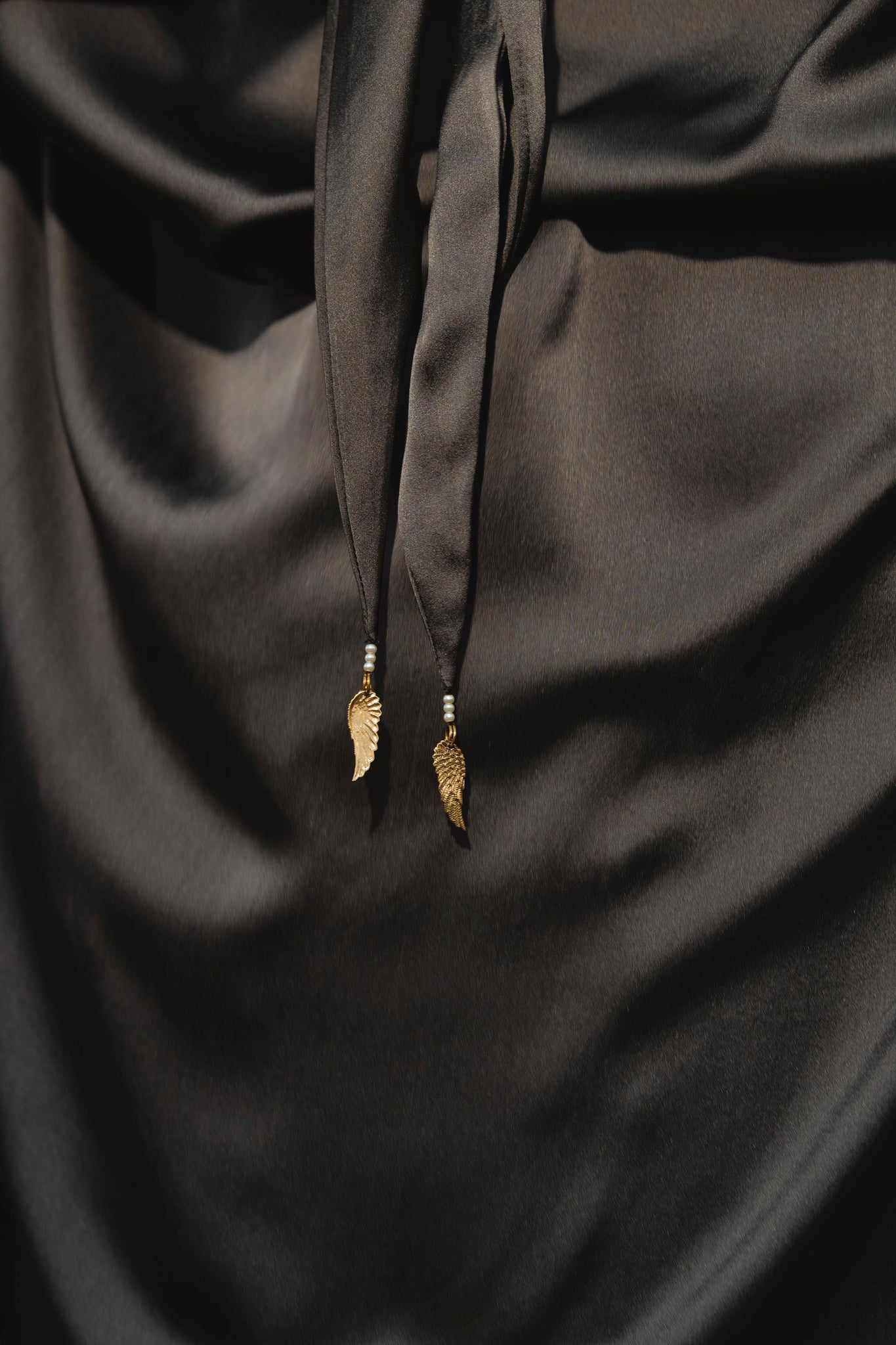 A close-up of smooth, shiny pure silk in asphalt gray on an Apsara Bra by AYA Sacred Wear, featuring two gold leaf-shaped charms on knotted ties at the center; handcrafted in Bali.