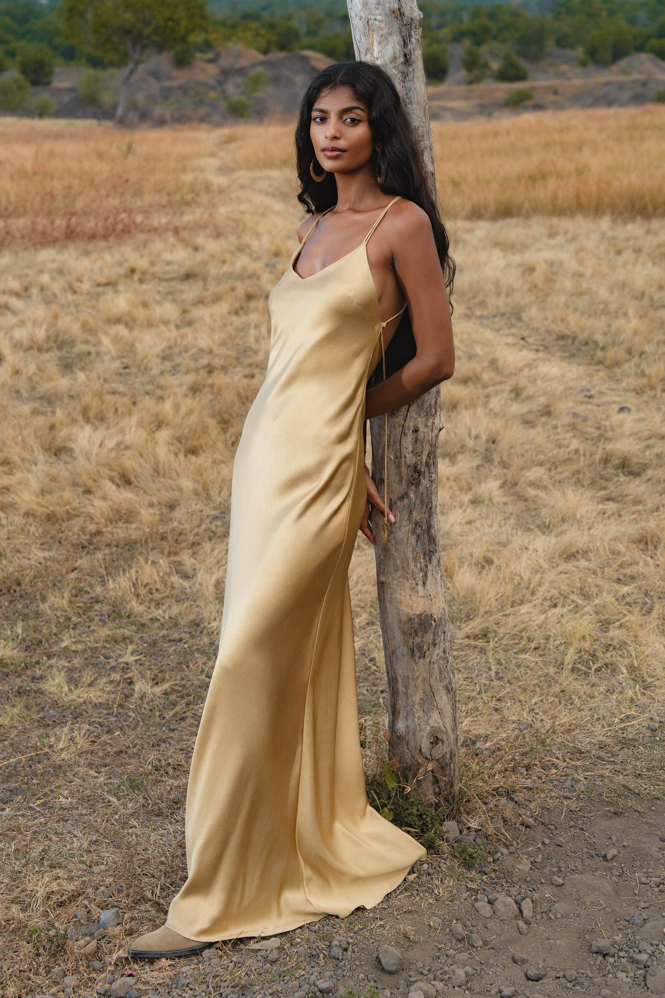 A woman in an AYA Sacred Wear Gold Silk Maxi Dress with a deep V-neck and backless design stands by a tree in a dry, grassy field with hills and trees in the background.