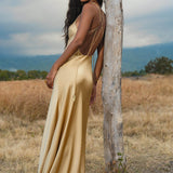 A woman in an AYA Sacred Wear Gold Silk Maxi Dress stands outdoors by a tree, showcasing the deep V-neck and backless design with thin straps. The evening gown glows against a grassy field with mountains and a cloudy sky behind her.