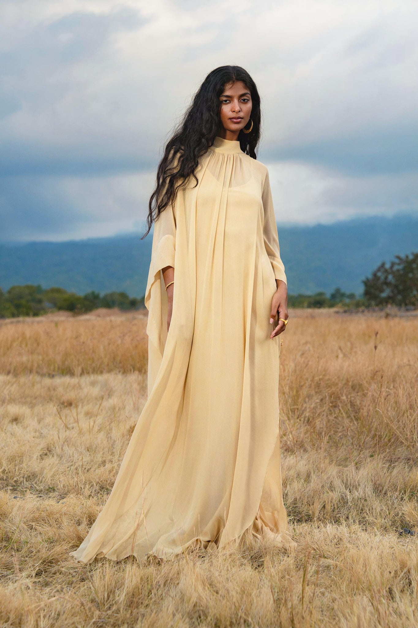 A woman with long dark hair stands in a dry grassy field, wearing the Mahadevi Dress Inka Gold Colour – Pure Silk Chiffon Goddess Gown by AYA Sacred Wear. Mountains and cloudy skies form a stunning backdrop.