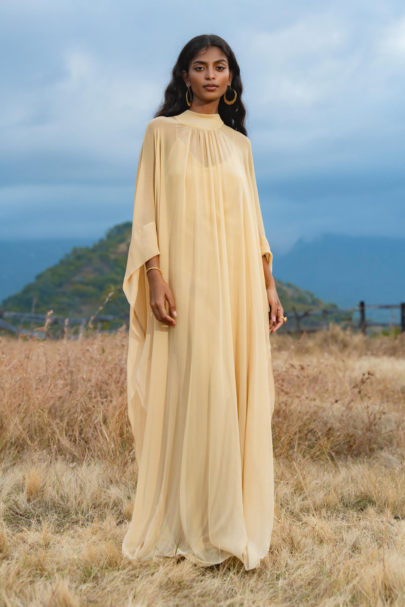 A woman with long dark hair and gold hoop earrings stands outdoors on dry grass, wearing the AYA Sacred Wear Mahadevi Dress Inka Gold Colour – a pure silk chiffon goddess gown, with mountains and cloudy skies in the background.