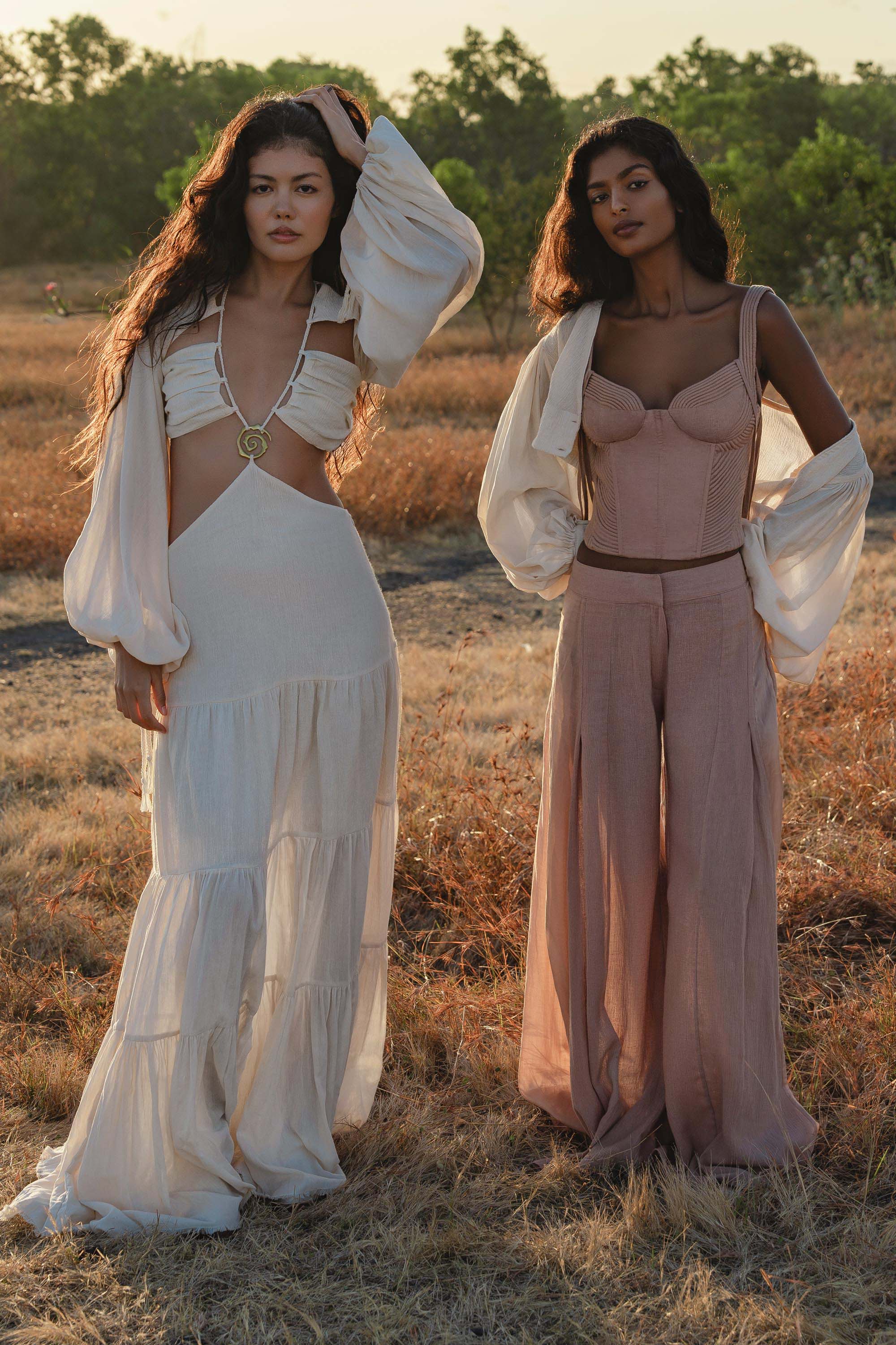 Two women stand on dry grass in boho style—one wearing the AYA Sacred Wear Samudra Dress in Ivory Colour, an organic bohemian dress with long sleeves and cutouts; the other wears a mauve bustier, wide pants, and a sheer cover-up. Both have wavy hair with brass spiral accents.