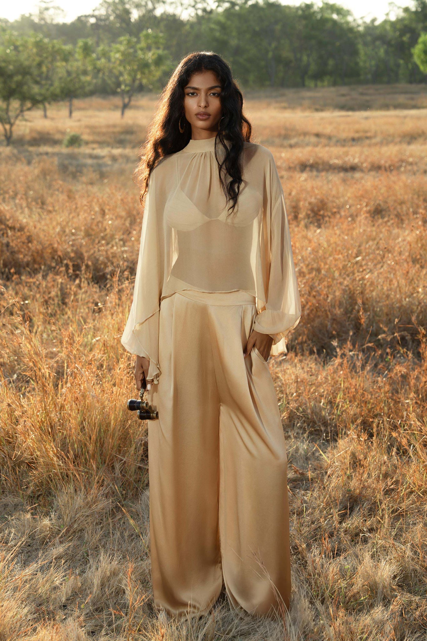 A woman with long, wavy hair stands in a grassy field, wearing AYA Sacred Wear’s Apsara Pants Inka Gold—pure silk flowing silhouette trousers—and a light gold blouse, holding a small camera as she gazes calmly ahead with trees in the background.