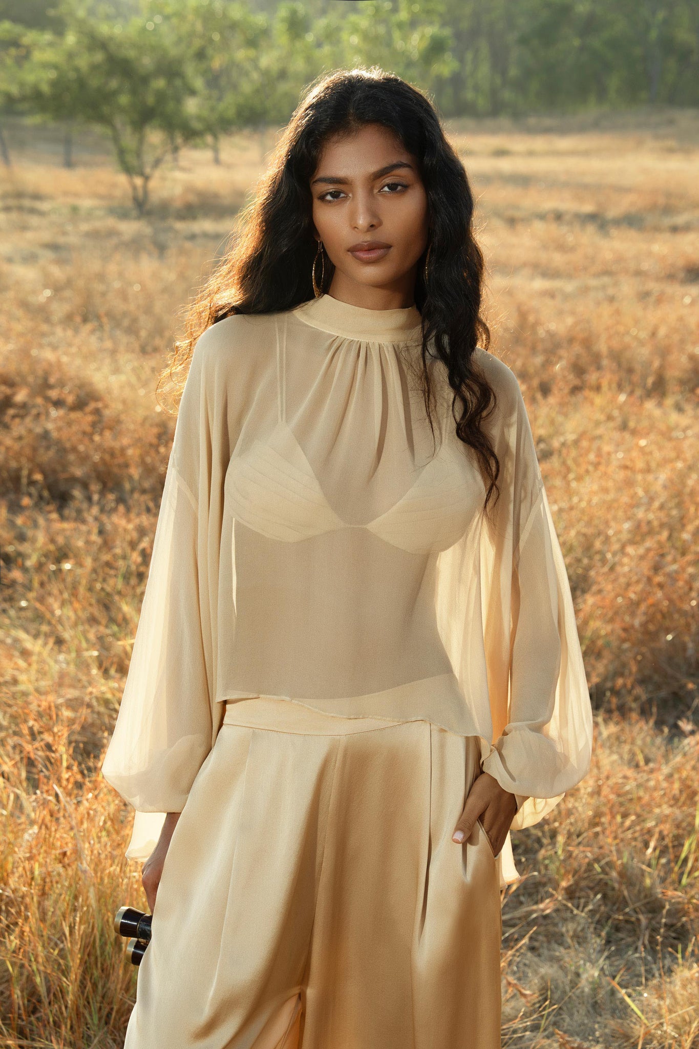 A woman with long wavy hair stands in a sunlit field, wearing a sheer beige blouse and AYA Sacred Wear's Apsara Pants Inka Gold—pure silk flowing silhouette trousers. She holds a small black object as trees appear in the background.