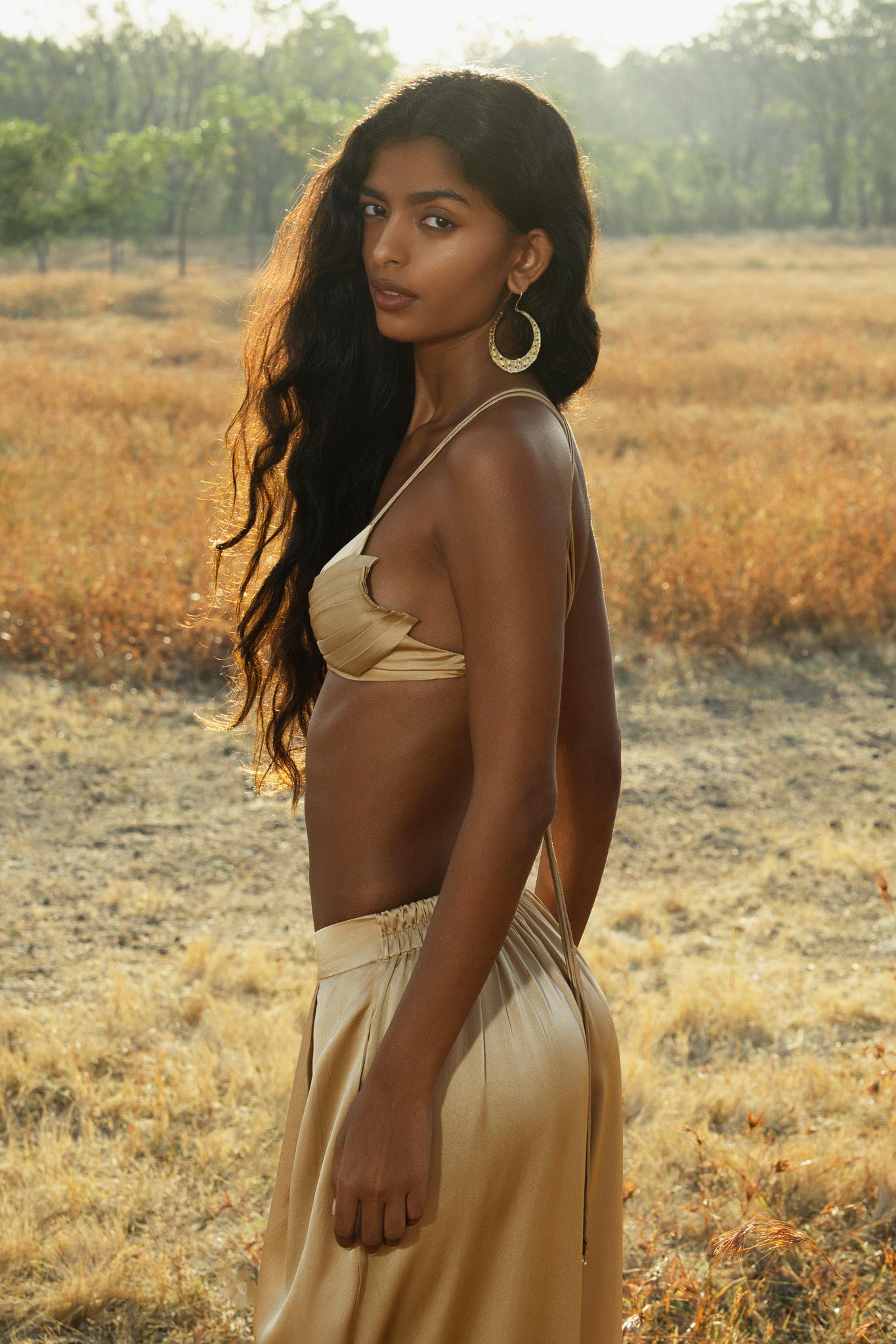 A woman with long wavy hair stands in a dry, grassy field, wearing the Butterfly Silk Bra in Inka Gold by AYA Sacred Wear—sculpted pure silk intimates handcrafted in Bali—while sunlight illuminates her face and body.