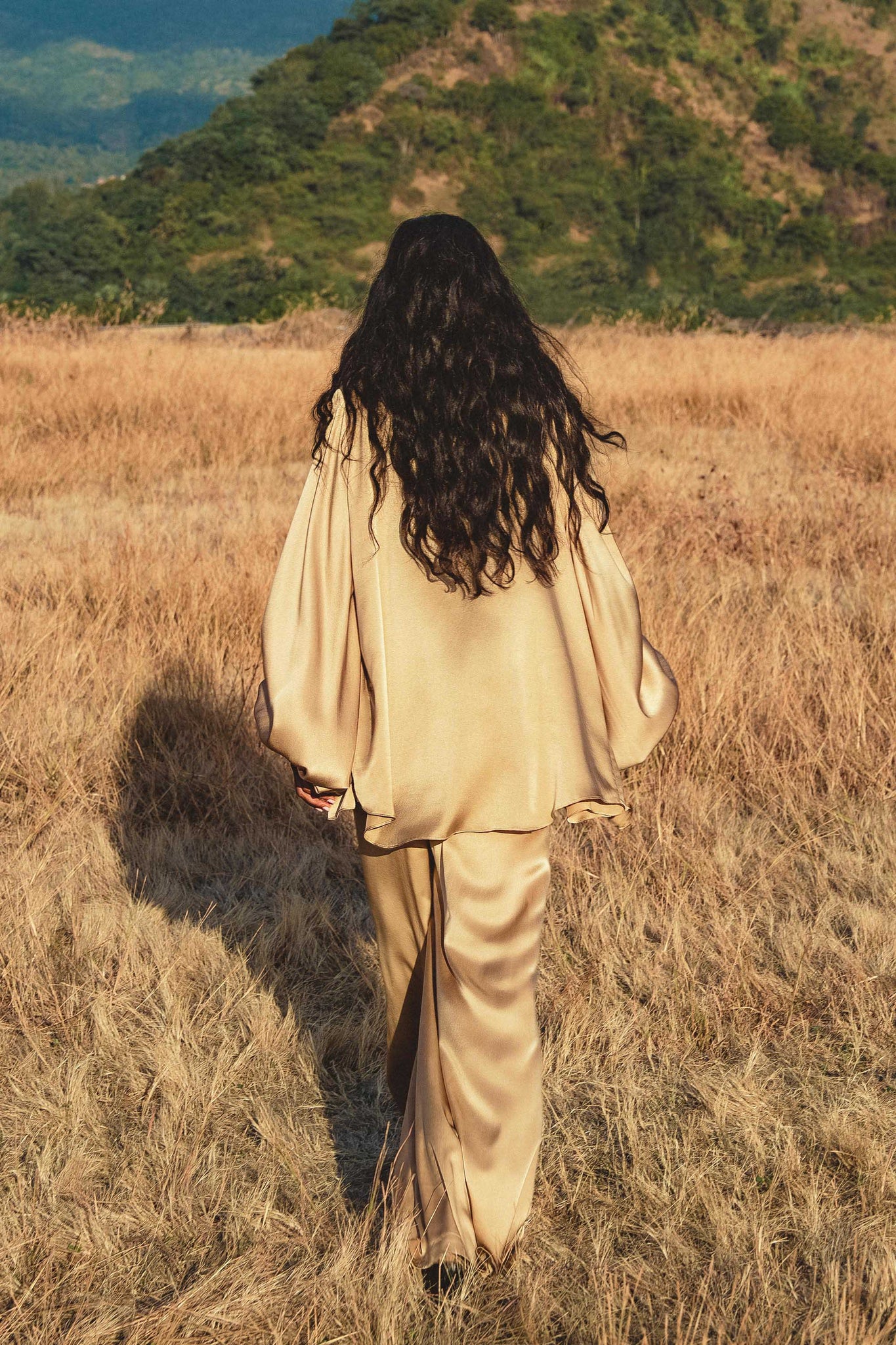 A person with long, wavy dark hair wearing the AYA Sacred Wear Apsara Kimono Blouse in Inka Gold—an elegant pure silk top—walks through a dry grassy field toward a tree-covered hillside under a clear sky.