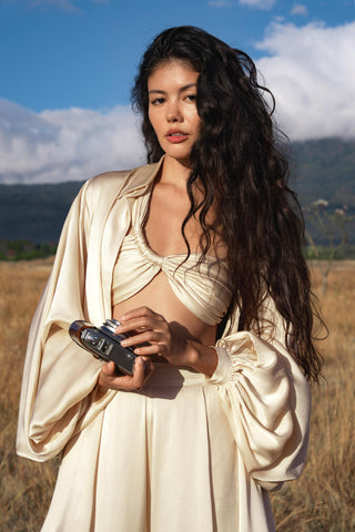 A woman with long, wavy dark hair stands in a grassy field, confidently holding a vintage camera. She wears the AYA Sacred Wear Apsara Bra Cream Colour — Pure Silk Transformative Top, with mountains and blue sky behind her.