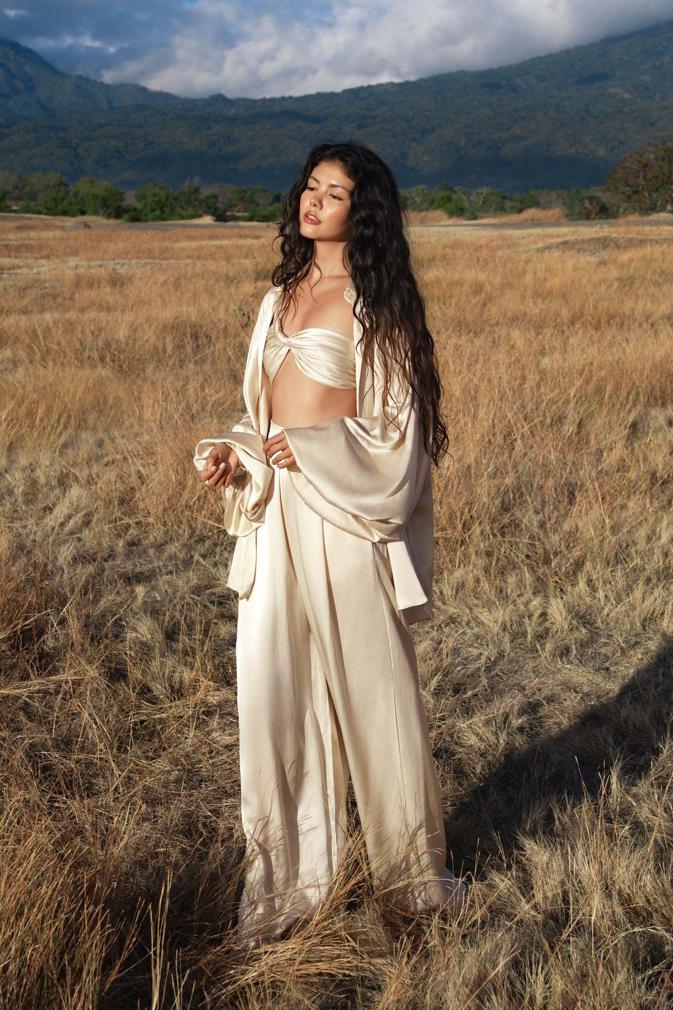 A woman with long, wavy hair stands in a dry grassy field, wearing the AYA Sacred Wear Apsara Kimono Blouse in cream—an elegant pure silk kimono top—paired with wide-legged pants. Mountains and a cloudy sky can be seen behind her.