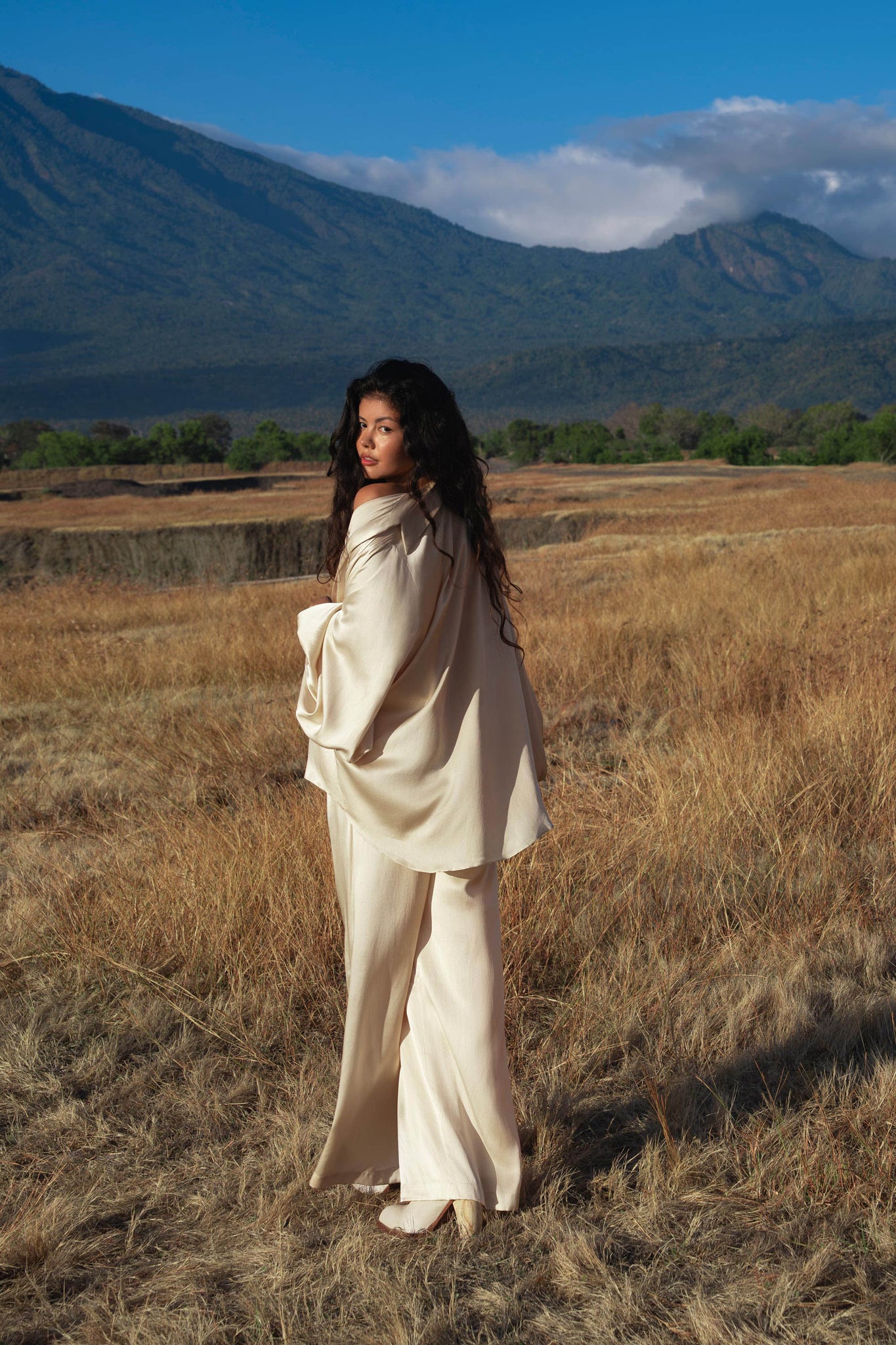 A woman with long dark hair wears the AYA Sacred Wear Apsara Kimono Blouse in cream, a pure silk kimono top, handcrafted in Bali, as she stands in a dry grassy field with green mountains and a partly cloudy blue sky behind her.