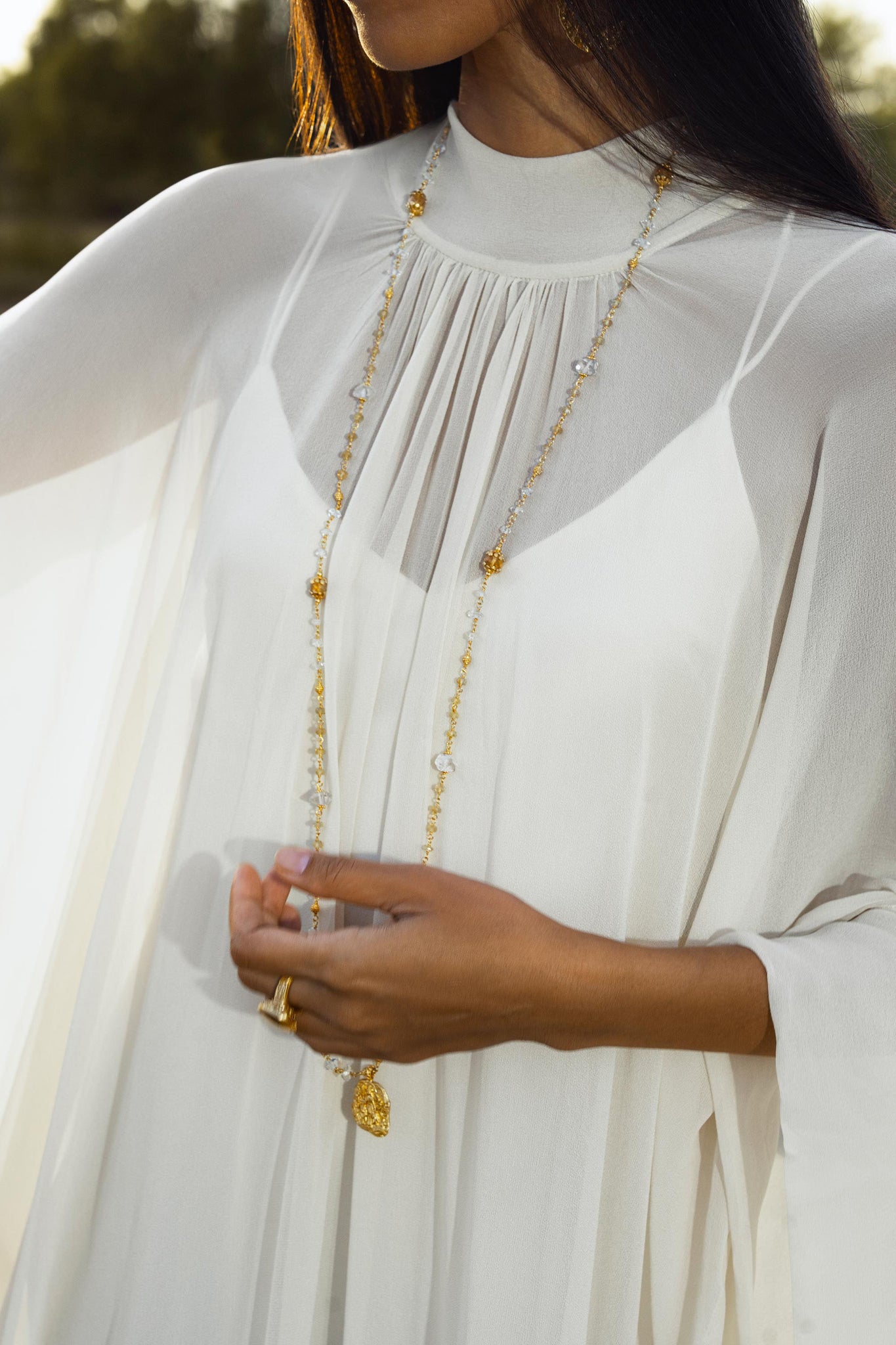 A woman outdoors wears the AYA Sacred Wear Mahadevi Dress Cream Colour – a pure silk chiffon goddess gown handcrafted in Bali – paired with a long gold charm necklace and matching rings, her face out of frame as she gently holds the necklace.