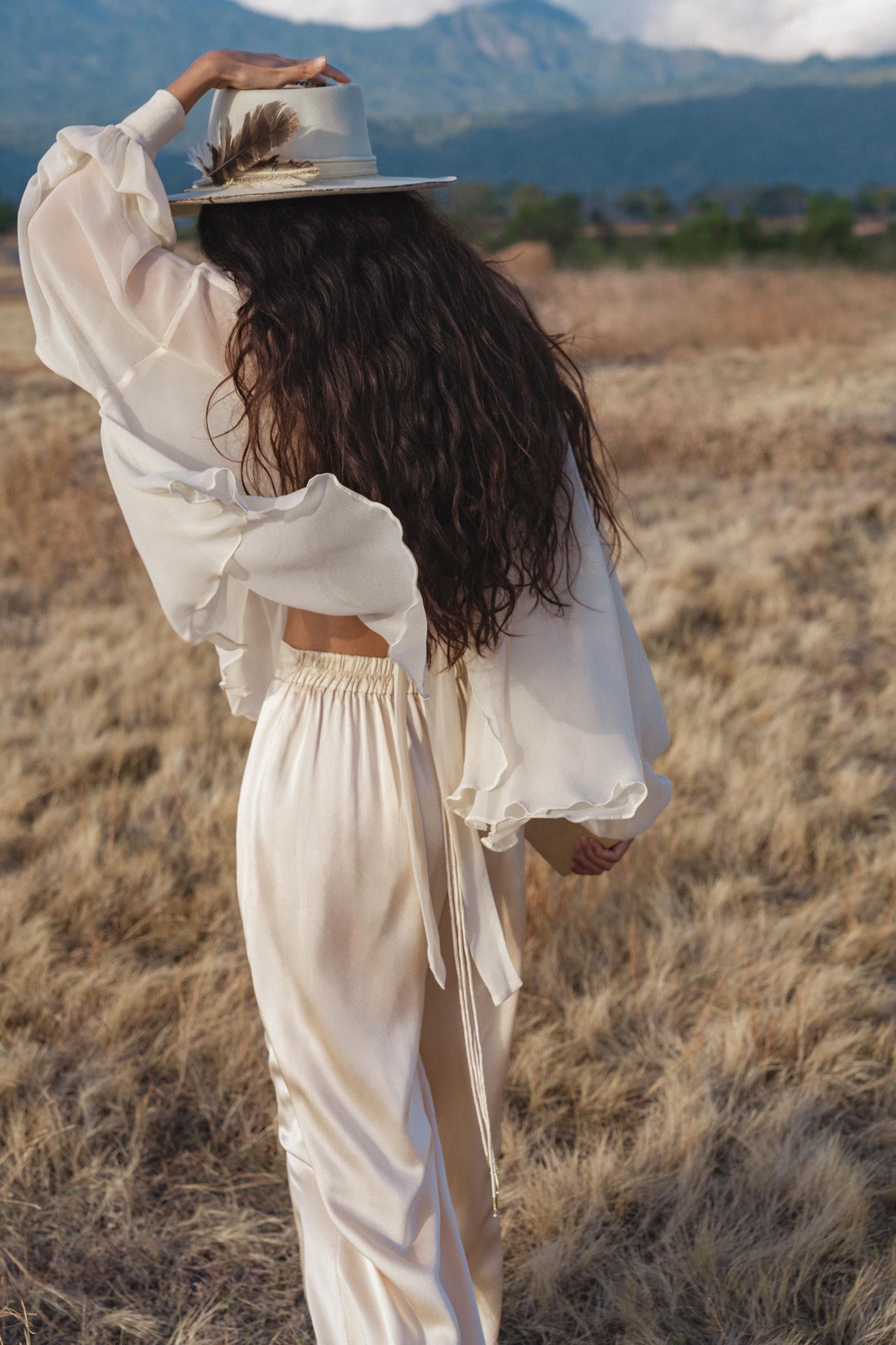 A woman with long wavy hair, seen from behind holding her wide-brim hat, walks through a dry grassy field in AYA Sacred Wear’s Apsara Pants Cream Colour—pure silk flowing silhouette trousers. Mountains rise in the distance.