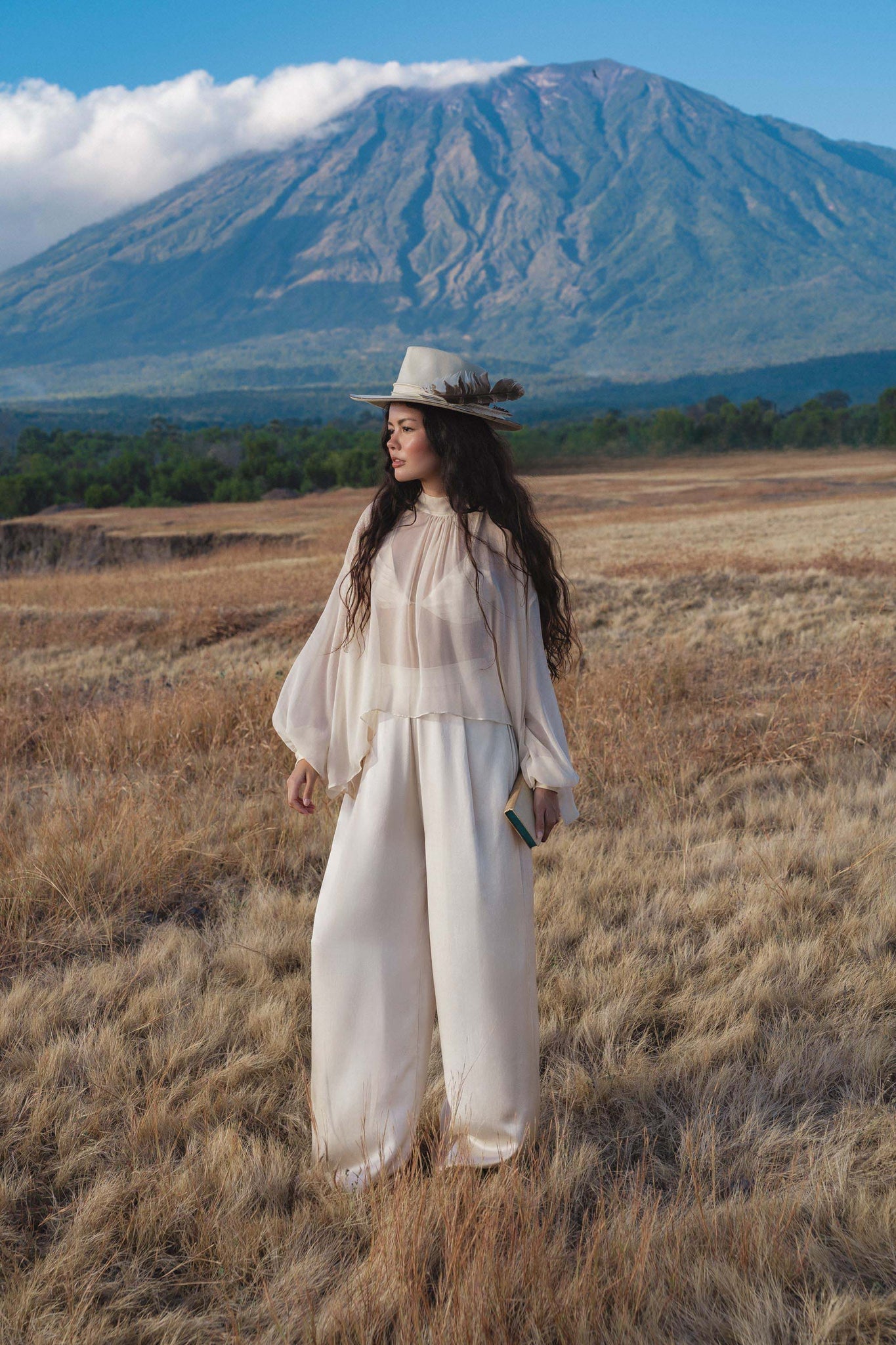 A woman wearing AYA Sacred Wear's Apsara Pants Cream Colour—pure silk flowing silhouette trousers—stands in a dry grassy field, holding a book, with a mountain in the background under a partly cloudy sky.