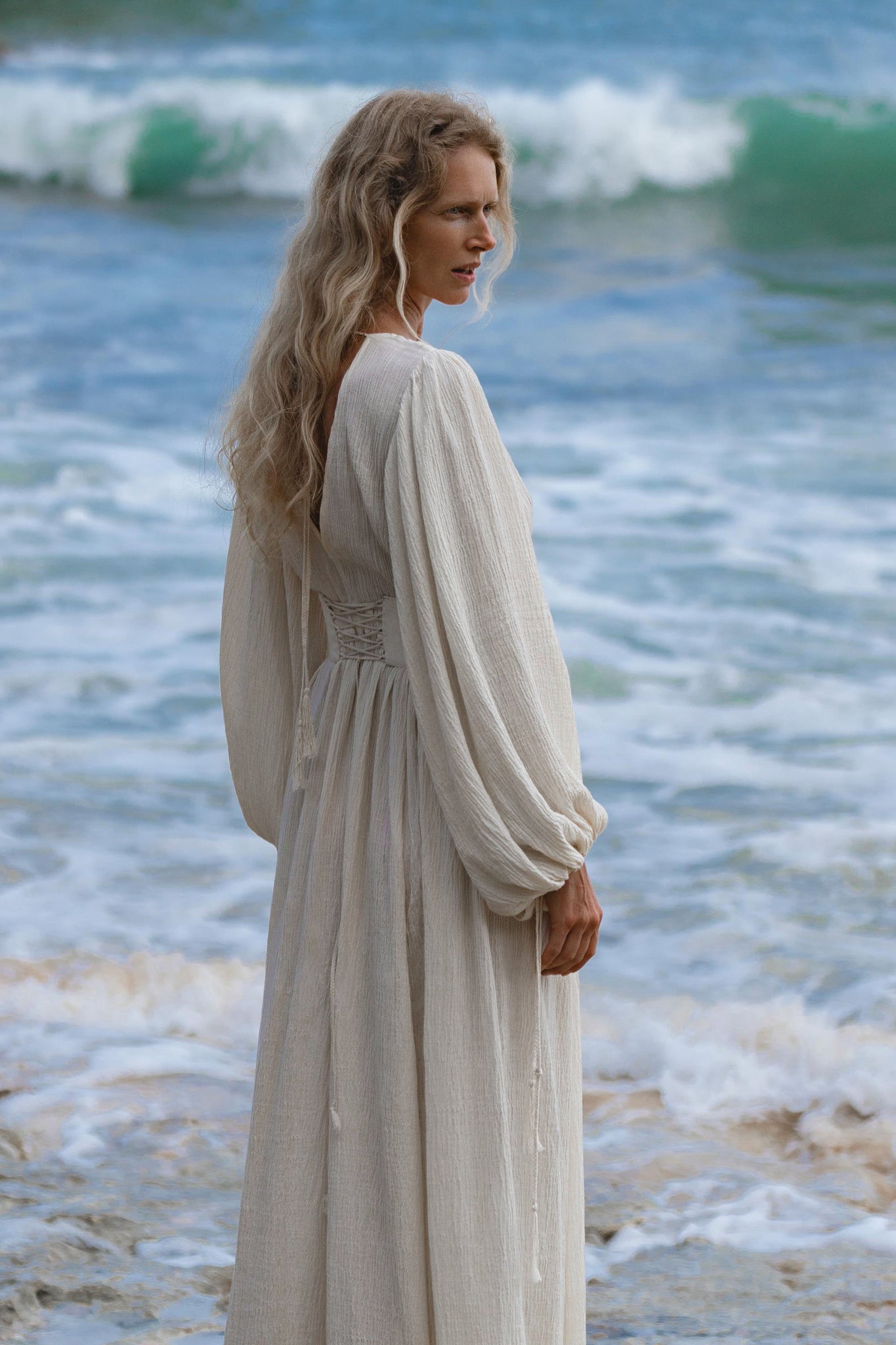 A woman with long, wavy blonde hair stands on the shore in the Tatva Dress Ivory Colour by AYA Sacred Wear, a sustainable bohemian cotton-linen dress with billowing sleeves, looking over her shoulder as gentle waves roll in behind her.