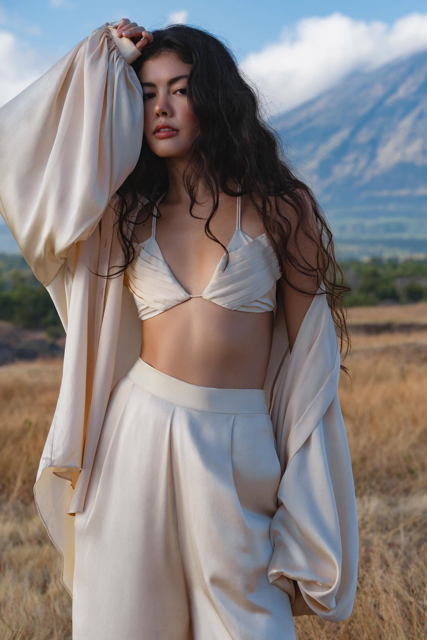 A woman with long, wavy dark hair stands in a dry field wearing the Butterfly Silk Bra in cream by AYA Sacred Wear, paired with wide-leg pants and an open shirt. She poses with one arm raised, mountains and blue sky in the background.