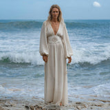 A person with long blonde hair stands on rocky sand near the ocean, wearing the Tatva Dress Ivory Colour—Sustainable Bohemian Dress by AYA Sacred Wear. Waves crash behind them as they gaze calmly at the camera under a cloudy blue sky.