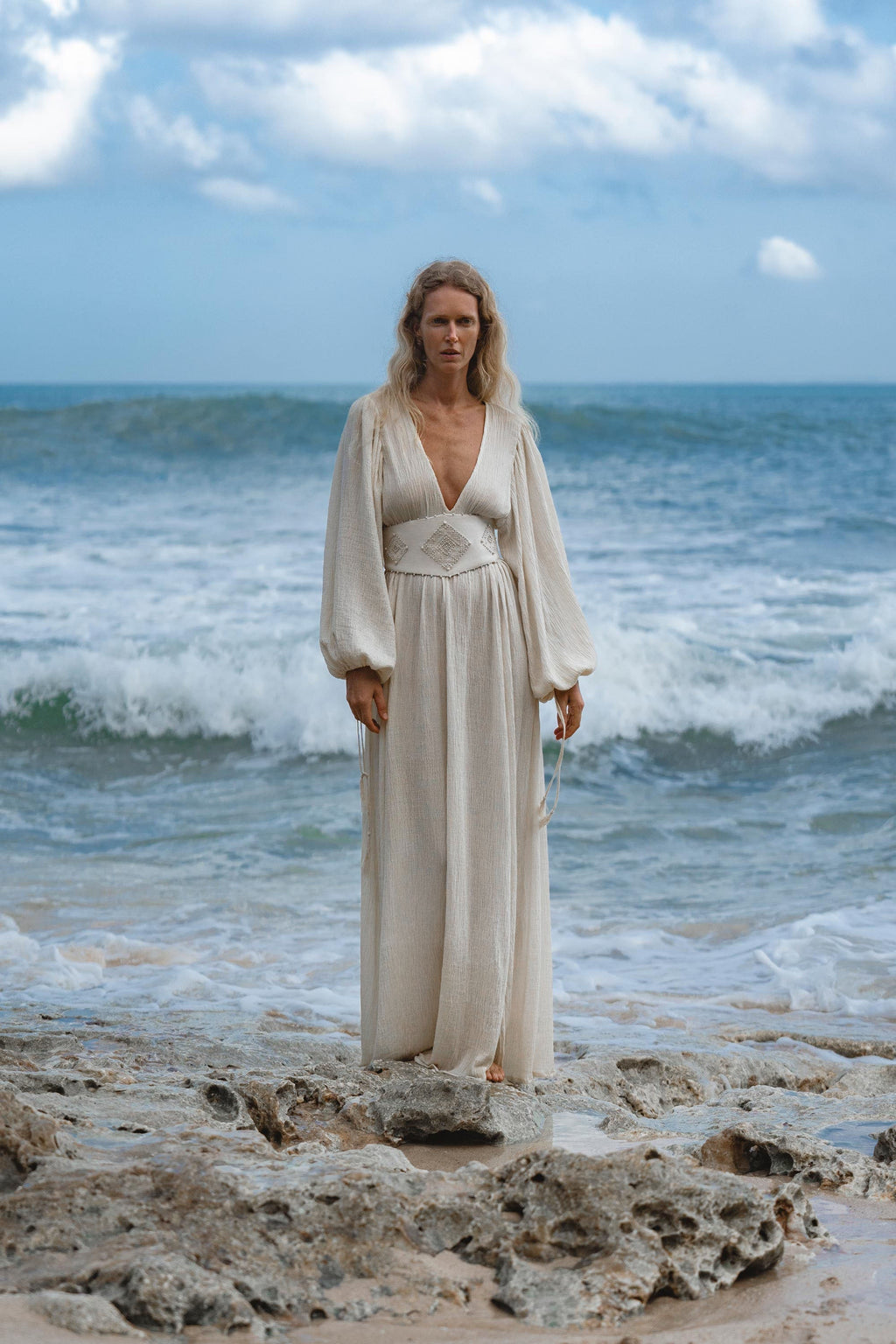 A person with long blonde hair stands on rocky sand near the ocean, wearing the Tatva Dress Ivory Colour—Sustainable Bohemian Dress by AYA Sacred Wear. Waves crash behind them as they gaze calmly at the camera under a cloudy blue sky.