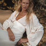A woman with long wavy blonde hair reclines on rocky sand, gazing calmly at the camera in the AYA Sacred Wear Tatva Dress Ivory Colour – a sustainable bohemian dress featuring textured cream fabric and a wide waistband.